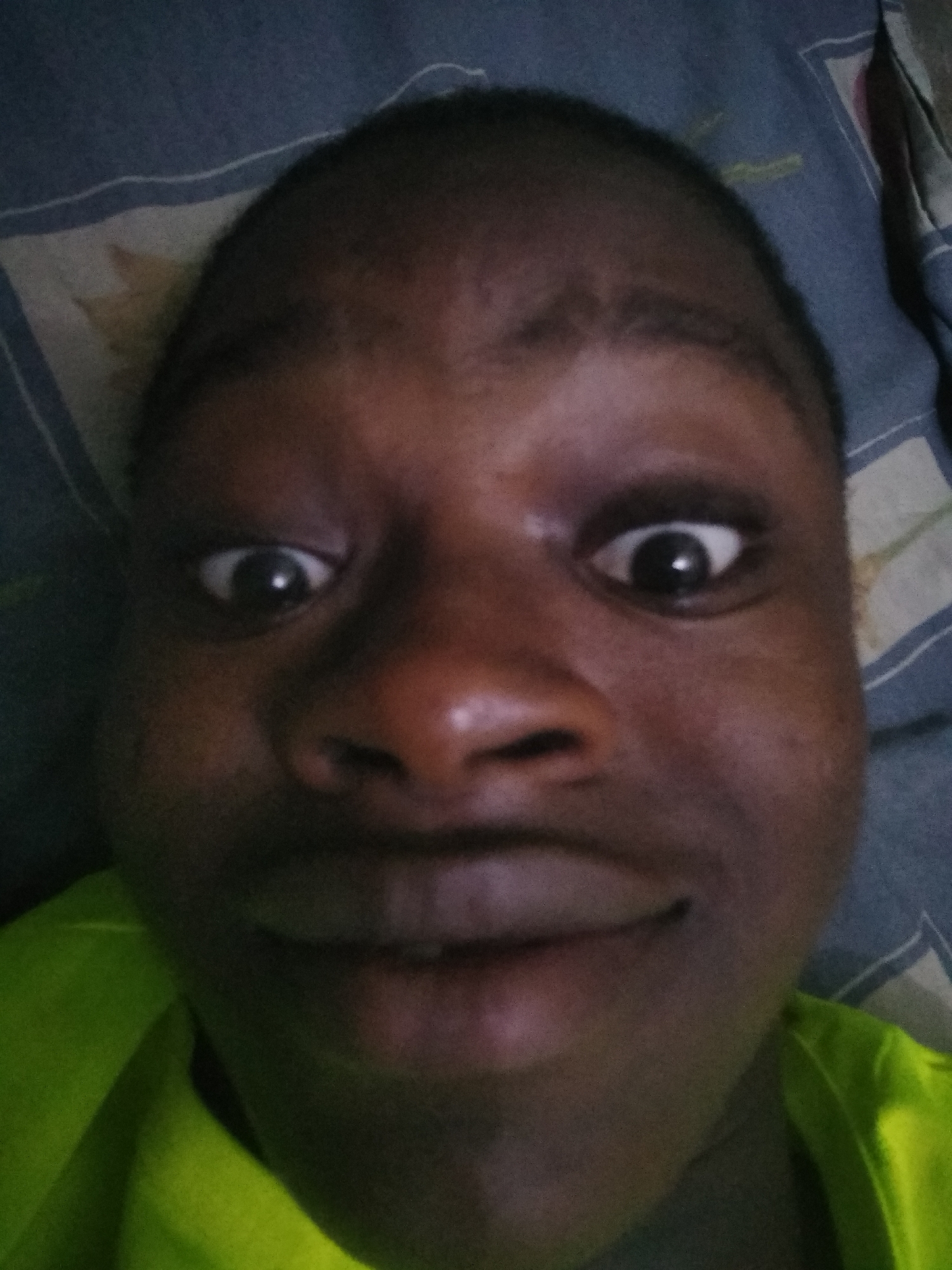 A close-up selfie of a dark-skinned person with wide eyes, a broad nose, and a closed-mouth smile. The person is wearing a bright green shirt. The background consists of a patterned blue and white fabric, possibly a bedsheet. The location in Shangisha, Nigeria, is not discernable from the image.