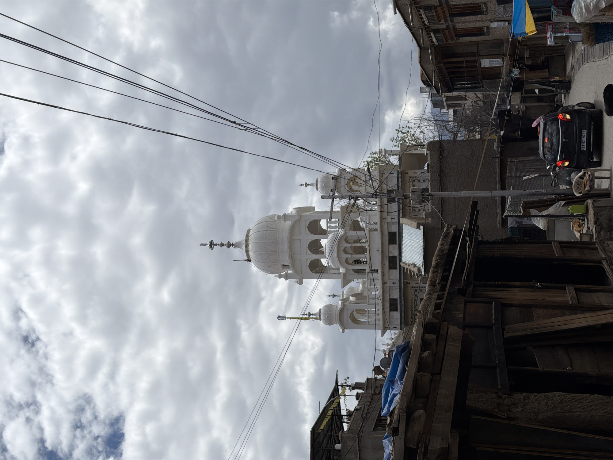 The image shows a street scene in the city of Leh, India, with a prominent white Sikh Gurudwara in the background. The Gurudwara features multiple domes and intricate architectural details. In the foreground, there are rustic wooden structures with corrugated tin roofs and blue tarpaulin. A black car with its brake lights on is parked on the street, and a white plastic chair is placed on the side. Electrical wires are strung across the sky, obscuring parts of the cloudy sky. The overall environment suggests a residential or commercial area in a mountainous region.