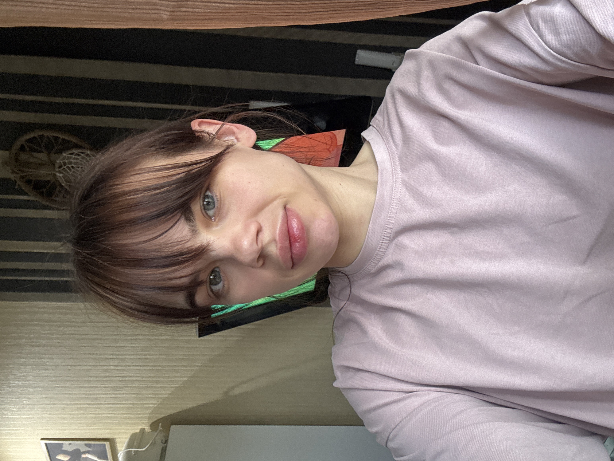 A woman with brown hair and bangs is looking directly at the camera. She's wearing a pink long-sleeved shirt. The background includes a wall with horizontal stripes and a woven object on the left. The location is Rostov-on-Don, Russia, but there are no details in the image confirming the exact place.