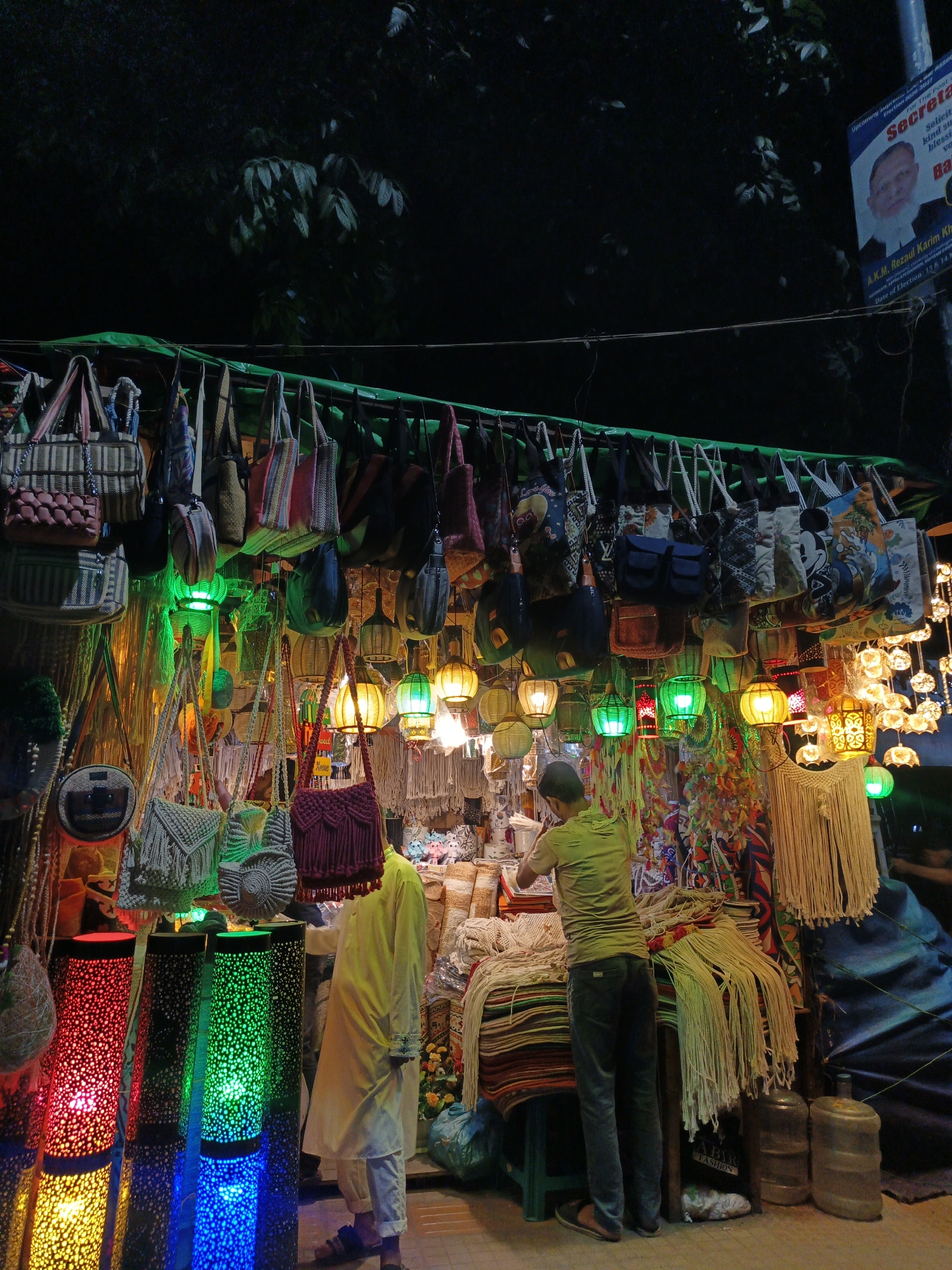 This media file captures a vibrant night scene at a market stall, likely in a South Asian country given the clothing of the individuals and the style of the stall. The stall is overflowing with a variety of items, primarily bags and decorative lamps, illuminated by colorful lights.
The main subjects are two people, seemingly a vendor and a customer, engaged in a transaction or display. One person, dressed in a light yellow shalwar kameez, stands to the right, partially obscured by the merchandise. The other, in a green t-shirt and dark jeans, is attending to items on the stall.
The stall is densely packed with a wide array of bags of different sizes, materials, and designs, hanging from a green canopy. Below the bags, an assortment of decorative lamps hangs, emitting colorful light in green, yellow, and red. There are also macrame hangings and woven textiles on display. In the foreground, tall cylindrical lamps are lit with red, green, and blue lights, casting a festive glow.
The scene is illuminated by the colorful lamps, creating a lively and somewhat chaotic atmosphere. The background is dark, suggesting it is nighttime. The presence of posters with text, including what appears to be election-related material with a candidate's photo, indicates a public space where such advertising might be displayed. The overall mood is one of bustling commerce and visual richness, characteristic of a night market. The text visible includes "Secretariat," "Election 2036-2017," and "A.K.M. Rezaul Karim Kh," suggesting a political context potentially related to the location or time.