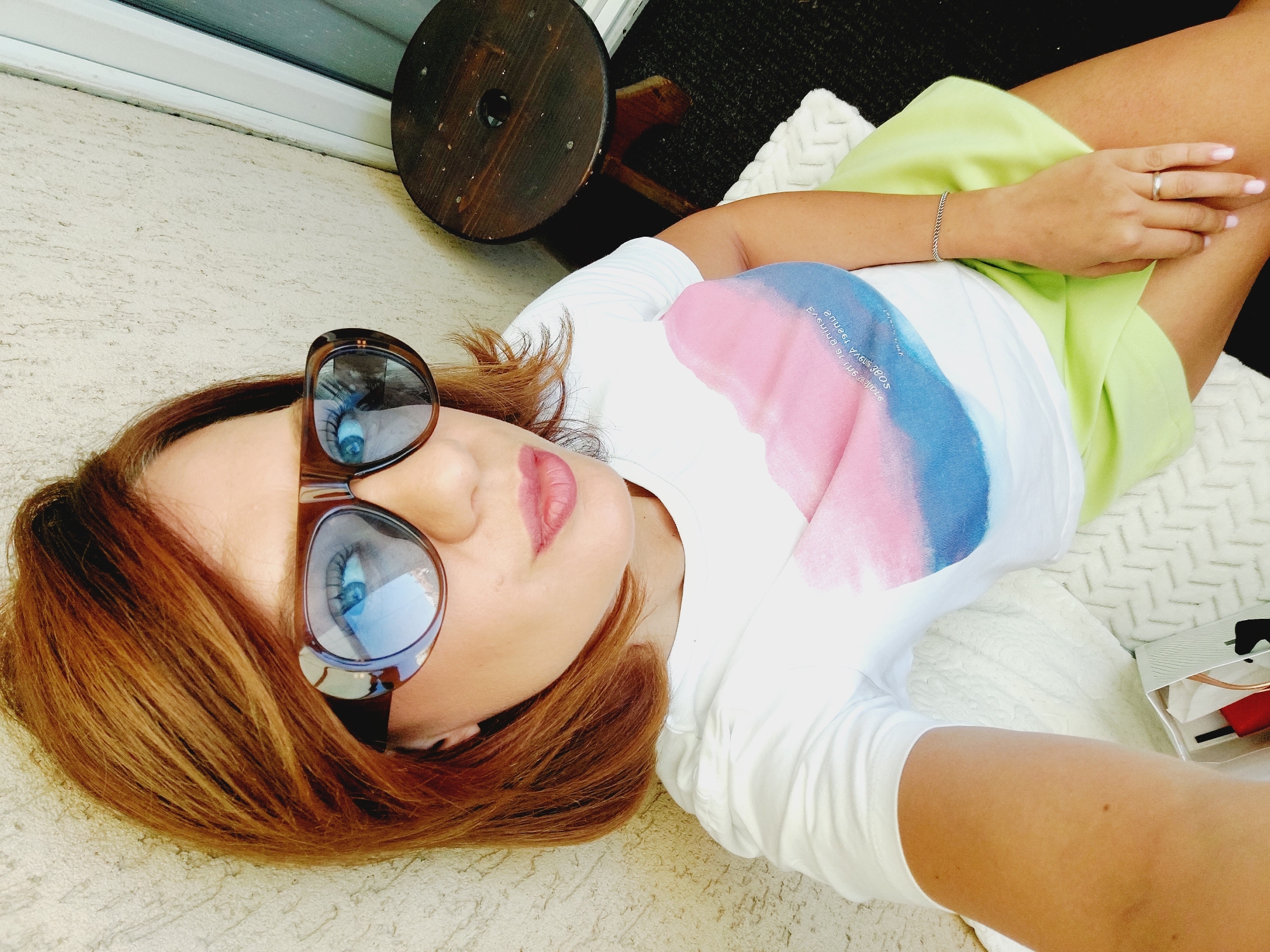 A light-skinned woman with shoulder-length auburn hair, wearing large brown-rimmed sunglasses, a white t-shirt with a pink and blue watercolor-style print, and a lime green skirt, is posing for a selfie. She has light pink lipstick and manicured nails. A silver bracelet is on her left wrist, and she wears a silver ring on her left ring finger. She is seated on a white patterned cushion outdoors, leaning against a textured cream-colored wall. To her right, a dark wooden stool with a circular top and three legs is visible, along with a sliver of a window frame and a dark floor covering. A white box containing items like red and black objects and what appears to be a white comb or brush is located in the lower-left corner of the frame. The overall scene suggests an outdoor balcony or patio space, possibly in Sofia, Bulgaria, given the context.