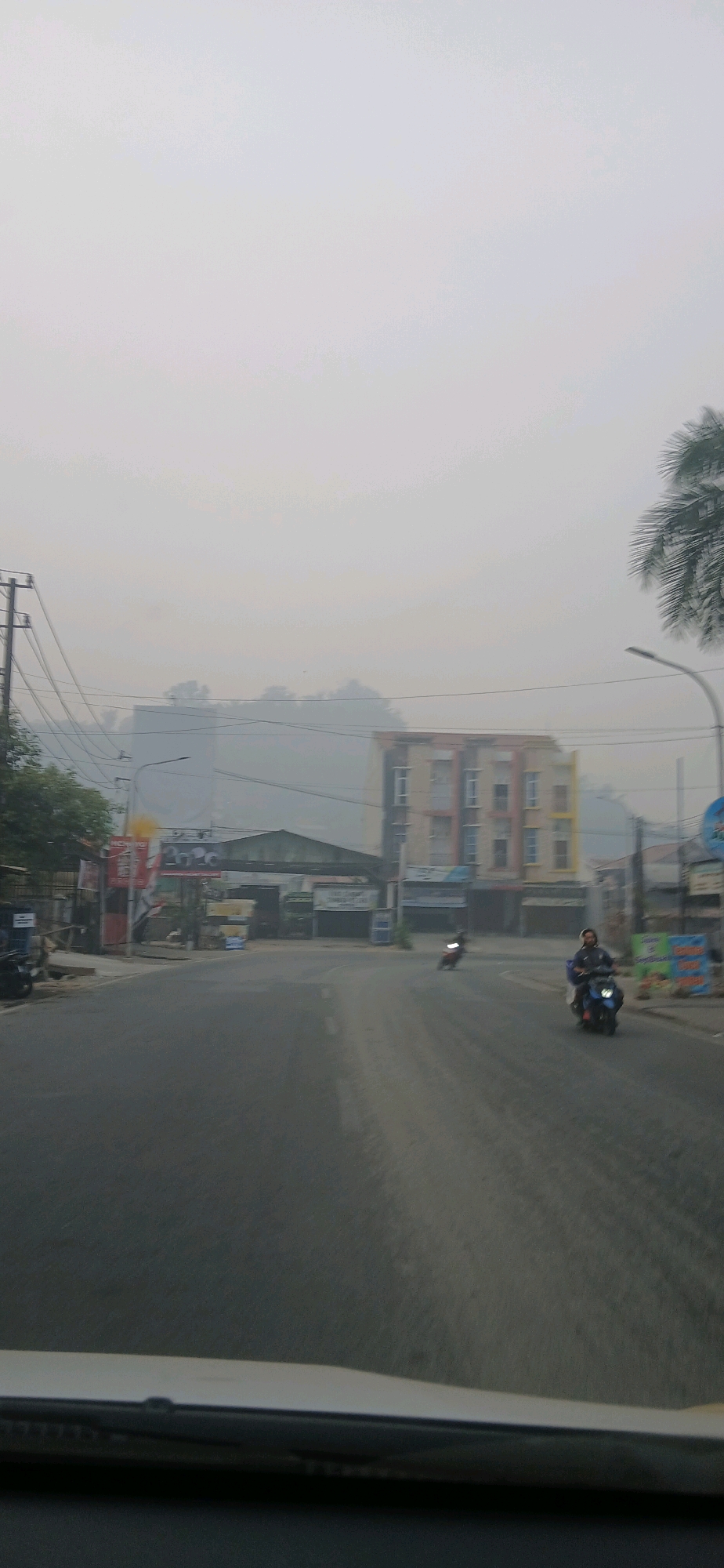 The image depicts a street scene in Samarinda, Indonesia, characterized by heavy haze or smog. In the foreground, the windshield of a vehicle is visible, suggesting the photo was taken from inside a car. The road ahead is paved and appears to be a two-lane street with some traffic.
On the right side of the road, a person is riding a dark-colored motorcycle. Further down the road on the right, there is a multi-story building with a light-colored facade and visible windows. A streetlamp is also present on this side. On the left side of the road, there are utility poles with overhead wires. Several buildings and structures are present, including what appears to be a covered entrance or a small building with a sloped roof. A red and yellow billboard is also visible on the left. In the distance, more buildings and trees are obscured by the thick haze, giving the impression of a dense urban environment with poor visibility. A palm tree frond is visible in the upper right corner, indicating a tropical or subtropical location. The sky is overcast and milky white due to the haze. The overall impression is one of a hazy day in an urban setting in Samarinda.