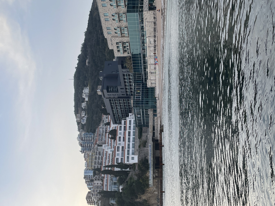 The image captures a coastal view in Budva, Montenegro, looking towards the shore from the water. The foreground consists of calm, dark water displaying subtle ripples, reflecting a diffused light from an overcast or hazy sky.
In the midground, a narrow strip of light-colored beach fronts a series of buildings. On the beach, several dark sun loungers are arranged, and a small group of three individuals is visible near three distinct colorful objects (red, blue, and yellow), possibly folded beach umbrellas or inflatables. Another individual is seated on the far right side of the beach. Directly behind the beach are modern, multi-story buildings; a large structure with extensive glass facades occupies the central portion, flanked by a cream-colored building with multiple arched balconies to its right. To the left of the central glass structure, a concrete pier or promenade extends over the water, supported by pillars, with lower, weathered structures underneath.
The background is dominated by a steep, densely forested hillside ascending from the coastline. Numerous multi-story residential and hotel buildings, exhibiting various architectural styles ranging from traditional white structures with terraced red roofs to contemporary designs with curved glass elements, are built into the slopes. Evergreen trees are prominent, particularly on the lower sections of the hill. A solitary communication tower or antenna is visible on the highest point of the forested hill. The sky is a uniform light grey, indicating soft, even lighting across the scene.