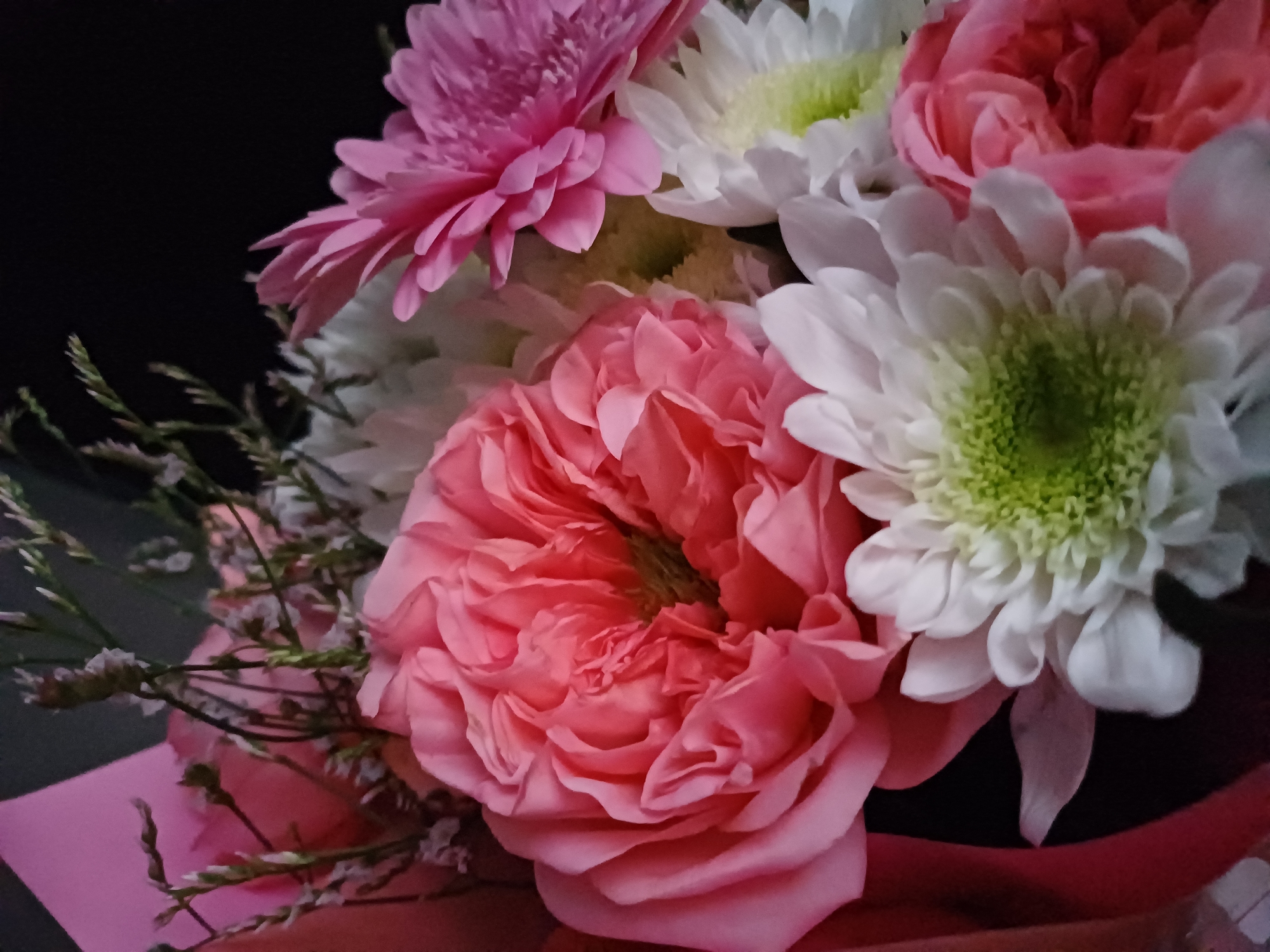 A close-up shot captures a bouquet of flowers against a dark background. The arrangement features several varieties, including a large, ruffled pink rose as the central focal point. To the right of the rose, a bright pink gerbera daisy with thin petals is visible. White chrysanthemums with green centers are interspersed throughout the bouquet, adding contrast. Delicate sprigs of greenery and small, pale flowers are also present, adding texture and depth to the floral composition. The lighting is soft, highlighting the soft textures and vibrant colors of the blossoms. The bouquet is partially wrapped in pink paper, adding another layer of color and visual interest. There are no people or identifiable landmarks visible in the image. The setting is indeterminable, but it appears to be an indoor arrangement.