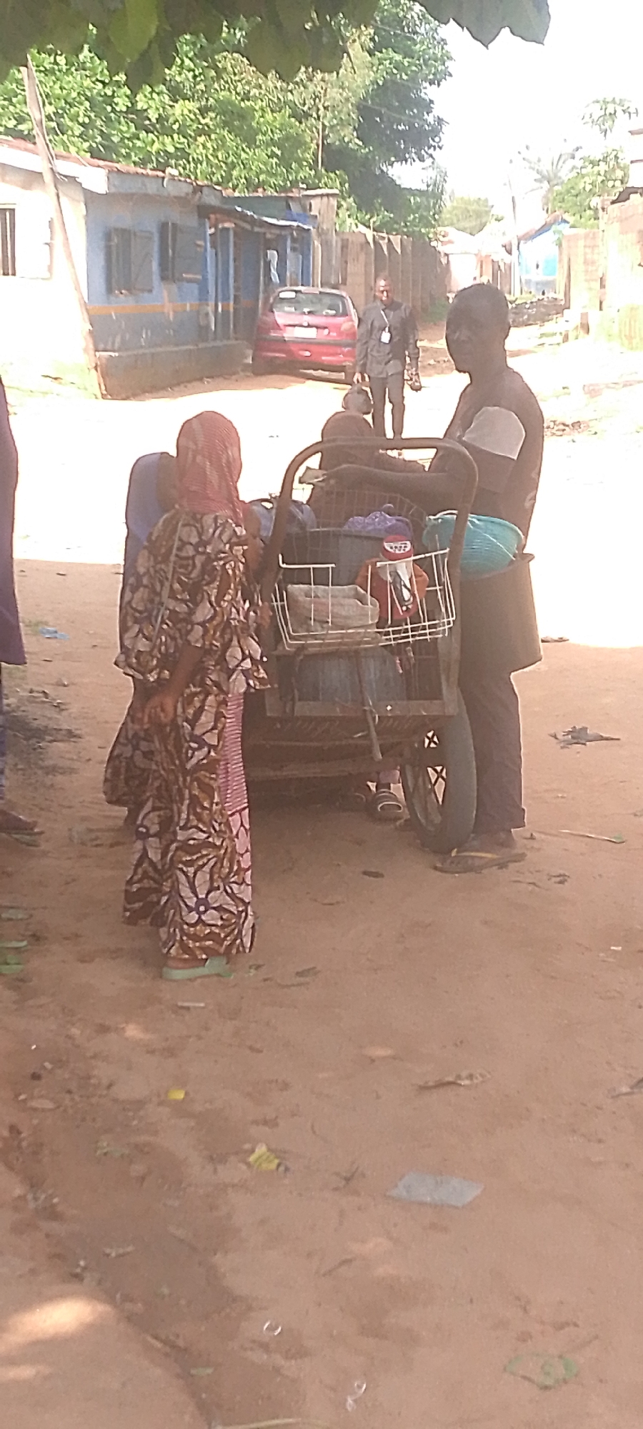 This media file captures a scene in Jalingo, Nigeria, depicting several individuals and objects in what appears to be a public outdoor setting. The overall impression is one of everyday life on a dusty street.
In the foreground, a woman wearing a colorful, patterned dress and a headscarf is visible from the waist down, facing towards a three-wheeled cart. She is wearing light-colored sandals. Next to her, partially obscured, is another person. The three-wheeled cart is laden with various items, including what looks like bags and fabric. The cart itself is metallic with a basket attachment in the front, and one of its wheels is clearly visible.
Behind the cart, two individuals are standing. One person, possibly a man, is wearing a dark shirt and pants and appears to be interacting with the cart. The other person, to the right, is seen from the chest up, looking towards the viewer. This individual is wearing a dark shirt and has a light-colored cloth around their neck or shoulder.
In the background, a red car is parked on the side of a street. Beyond the car, there are buildings with corrugated metal roofs and faded paint, suggestive of a modest urban or semi-urban environment. The buildings appear to be in a state of disrepair or have an aged look. Wooden fences and some trees with green foliage are also present, indicating a somewhat rural or less developed setting. The ground is covered in dirt and appears dry and dusty.
The lighting suggests it is daytime, likely morning or afternoon, with sunlight casting shadows and illuminating the scene. There is no obvious indication of any particular event or ceremony taking place; rather, it seems to be a candid snapshot of daily activity. The overall mood appears neutral, focusing on the people and their surroundings. No visible text is discernible in the image.