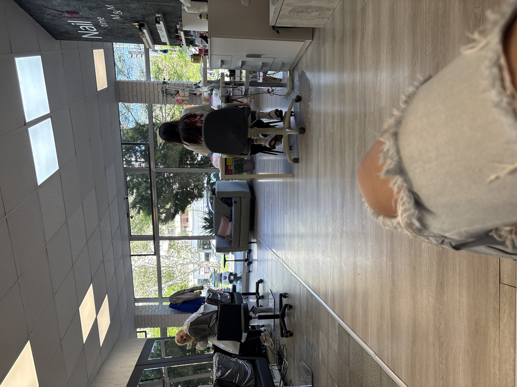 The image, correctly oriented, shows the interior of a modern nail or beauty salon, well-lit by natural light streaming through large windows spanning one side of the room. The floor is covered in light wood-look laminate.
In the foreground, the shoulder and arm of a person, likely the photographer, wearing light-colored, distressed denim, are visible and out of focus. Further into the salon, two customers are seated in black chairs with patterned backs, facing away from the camera. One customer, with dark hair, is at a station with white desks and salon products. A blonde-haired stylist, wearing a grey and white smock, stands facing the camera while attending to a client at another station. A third customer is also seated further down the room, facing away.
Salon equipment, including white desks, wheeled chairs, and shelves stocked with beauty products, is arranged throughout the space. A grey sofa with a pink cushion serves as a waiting area along the windowed wall. Through the windows, green trees, other buildings, and a white scooter are visible, suggesting a sunny daytime setting. On a dark wall, a sign reads "Nail time M&M". The overall impression is a busy, functional salon in Plovdiv, Bulgaria.