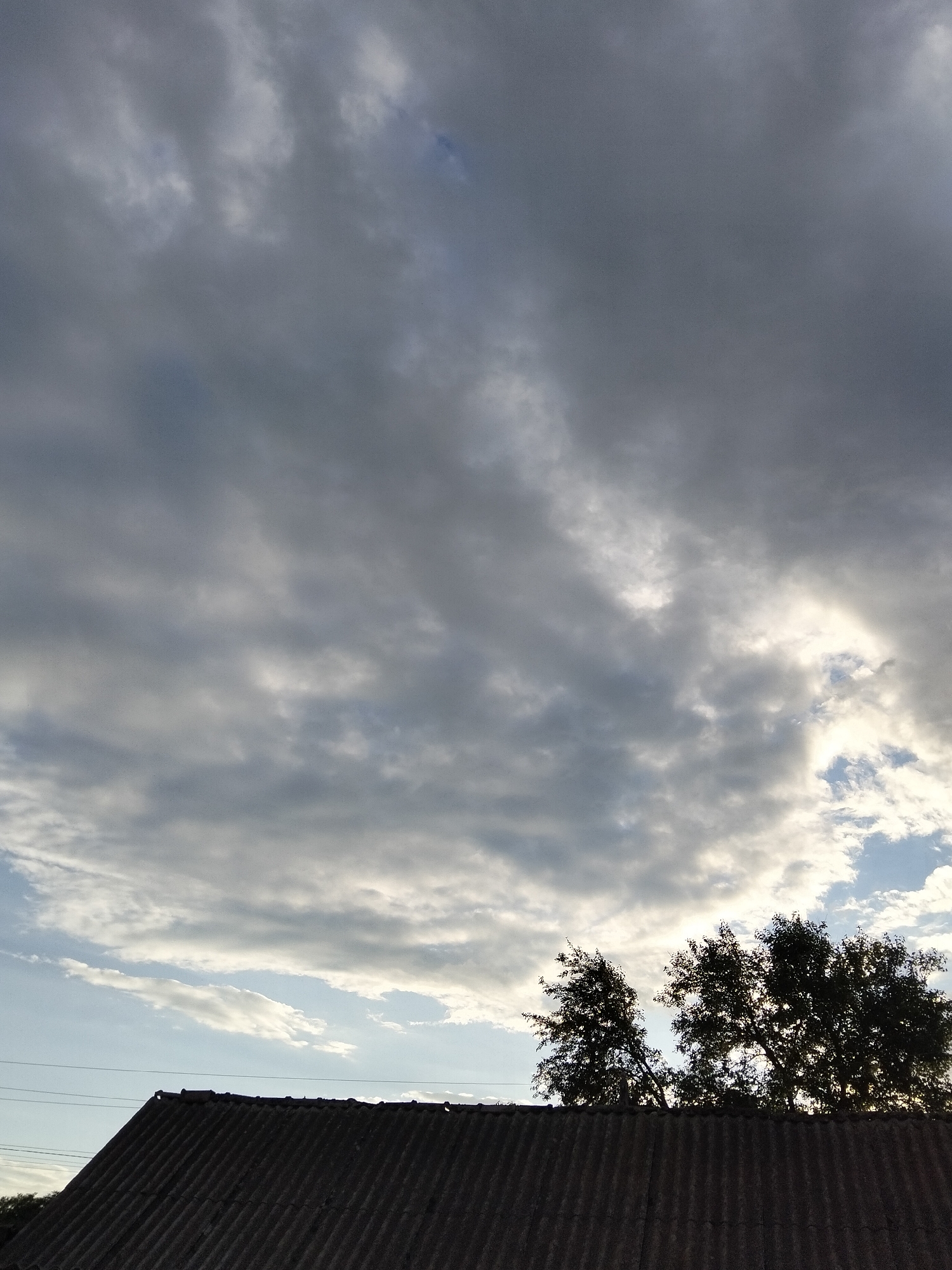 A rooftop in Ustyvytsia, Ukraine, is shown under a cloudy sky. The rooftop is made of corrugated material and slopes downwards from right to left. Behind the rooftop, silhouetted trees with dense foliage are visible against the sky. The sky is filled with a mix of dark gray and lighter gray clouds, with some patches of bright sky peeking through, particularly on the right side. The overall lighting suggests it is either early morning or late afternoon, with the sun obscured by the clouds. No people or specific actions are depicted in the image.