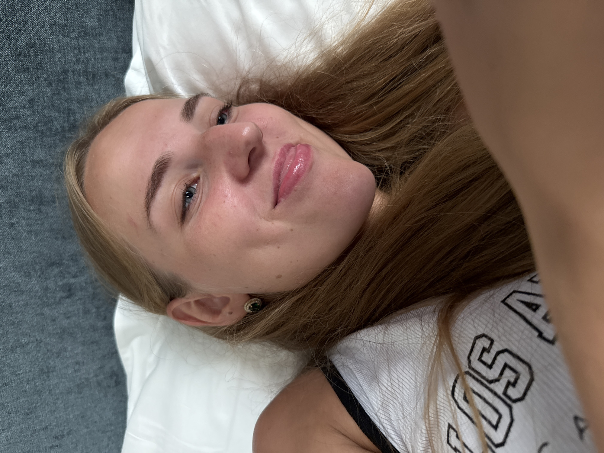 The image shows a close-up of a young woman with blonde hair lying on a white pillow. She is wearing a white tank top with black lettering that appears to say "LOS A". Her head is resting on a dark gray textured surface, likely a headboard or wall. She has light blue eyes, fair skin with some freckles, and is wearing a small green earring. Her lips are slightly parted and appear to have a glossy finish. The image is taken from a slightly high angle. There is no discernible city in Trieste, Italy, visible in the image.