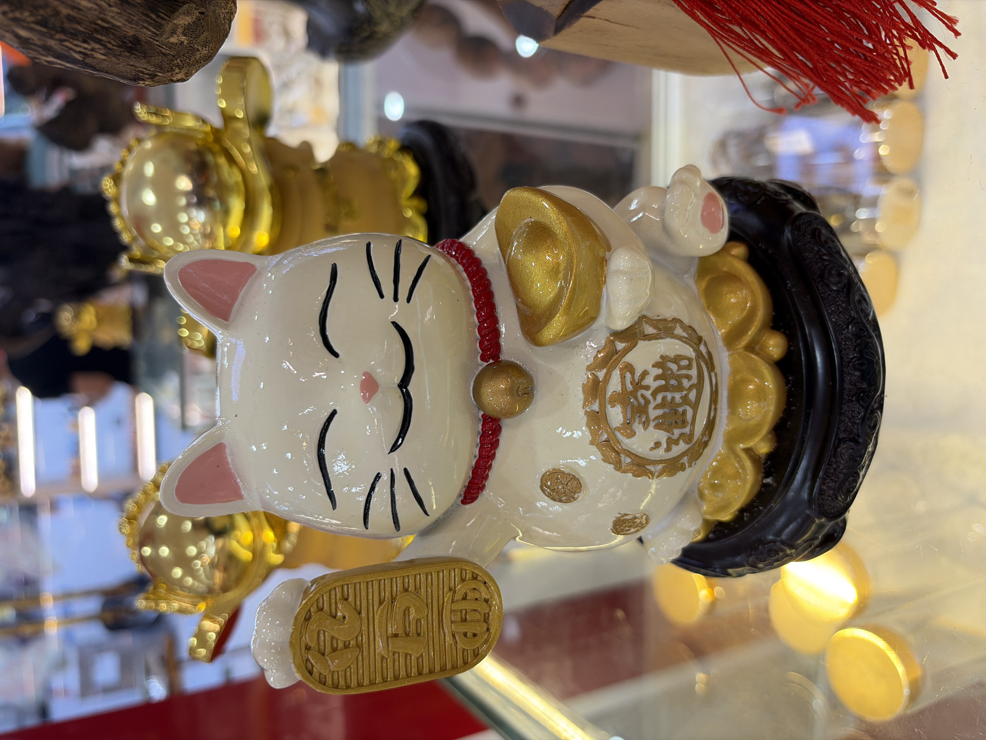 A close-up, slightly elevated, eye-level shot shows a white ceramic Maneki-neko (beckoning cat) figurine prominently displayed on a dark, textured base. The cat is facing forward with its eyes closed in a smile, its left paw raised and holding a gold-colored coin with Chinese characters. Its right paw is holding a gold-colored ingot. The cat wears a red beaded necklace with a gold bell. Circular gold embellishments are visible on its belly, and a circular gold emblem with Chinese characters is on its chest. The figurine is positioned on a glass surface, which reflects bright yellow light sources and some other circular objects.
In the blurred background, several other gold-colored figurines are visible, one of which appears to be a similar cat figurine. Further back, shelves and displays are faintly discernible, suggesting the location is a shop or market. A red tassel hangs to the right of the main figurine, partially obscuring a wooden object. The overall setting indicates a commercial environment selling decorative or auspicious items, likely in Đà Nẵng, Vietnam, given the cultural context of such figurines.