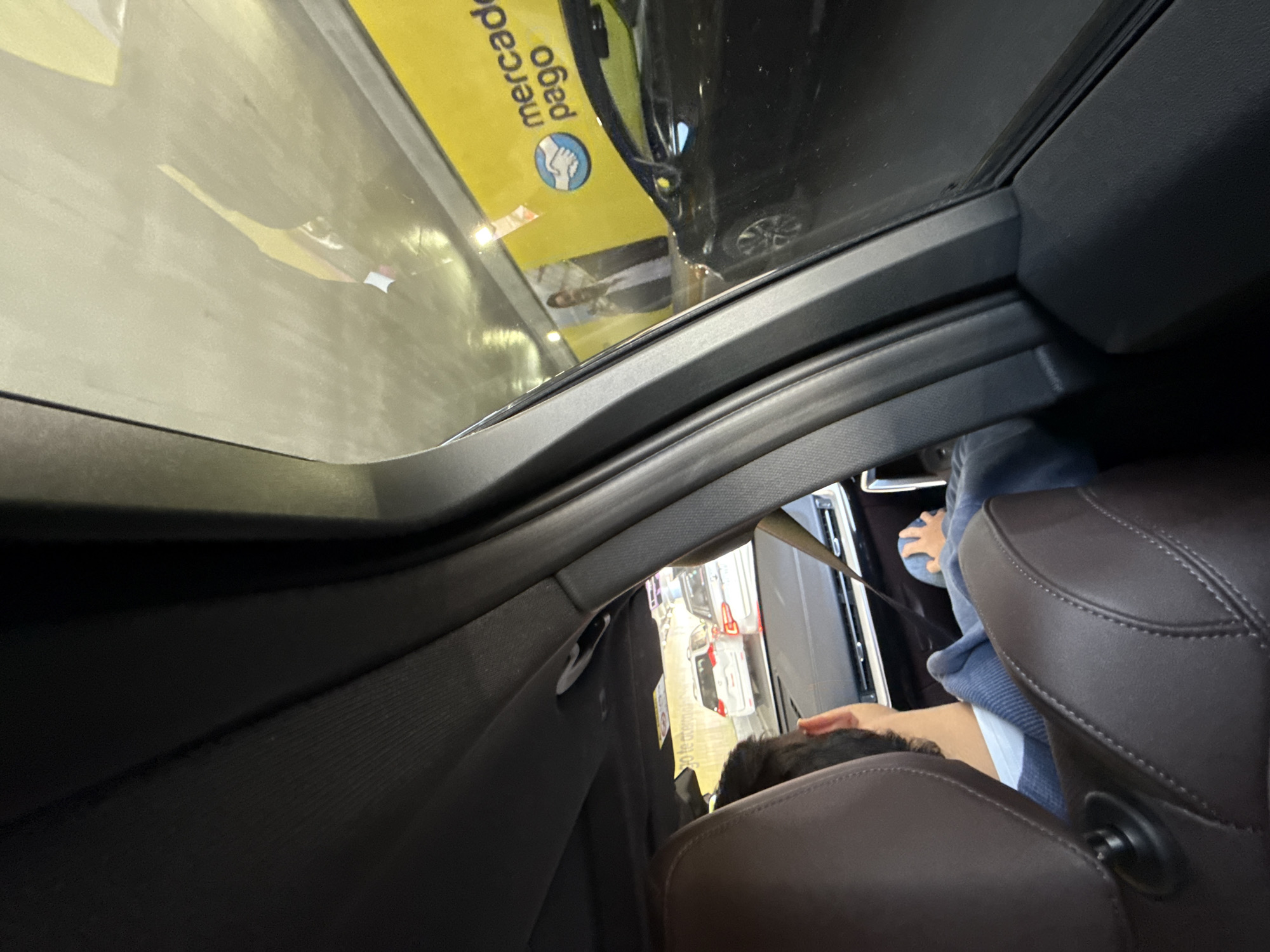 Here is a detailed description of the image:
The image is taken from inside a vehicle, looking upwards through an open sunroof. The frame of the sunroof and interior components of the car are visible.
Outside the vehicle, a Mercado Pago sign is prominently displayed, and other cars are parked or driving.
Inside the car, at least two people are present. In the foreground, the back of the head of a person with dark hair is visible. In the background, there is a person wearing a blue shirt or sweater. The person is sitting in the back seat.
The location appears to be Mexico City, Mexico due to the Mercado Pago sign.