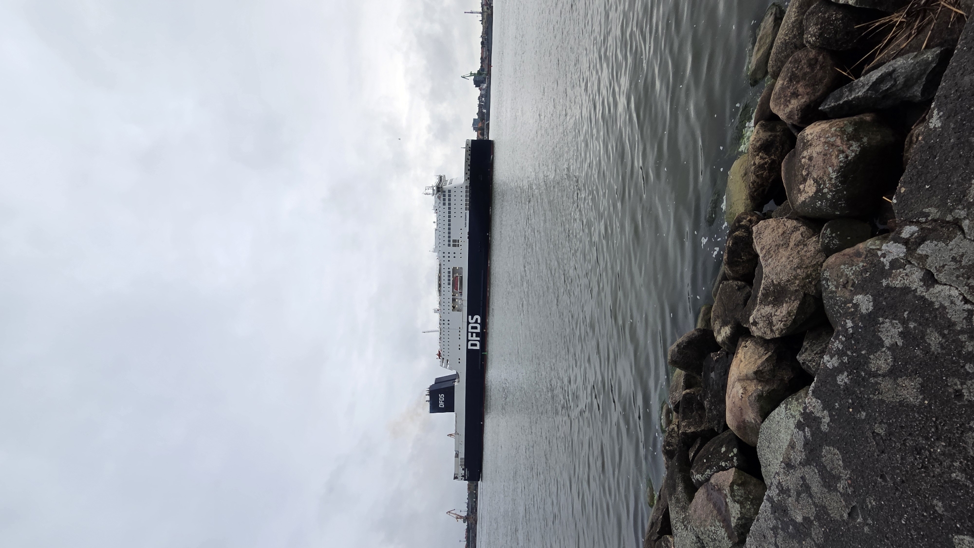 The media file shows a large ferryboat docked in a body of water. The ferry is dark with white upper decks and the word "DFDS" is visible on its side. The ferry appears to be a passenger and vehicle ferry. It is a cloudy day and the water is calm. The foreground of the image is filled with rocks, suggesting the photo was taken from a pier or shoreline. In the background, there are hints of a city or industrial area with cranes and buildings visible. The image is oriented vertically. There is no visible text other than the "DFDS" logo on the ferry.