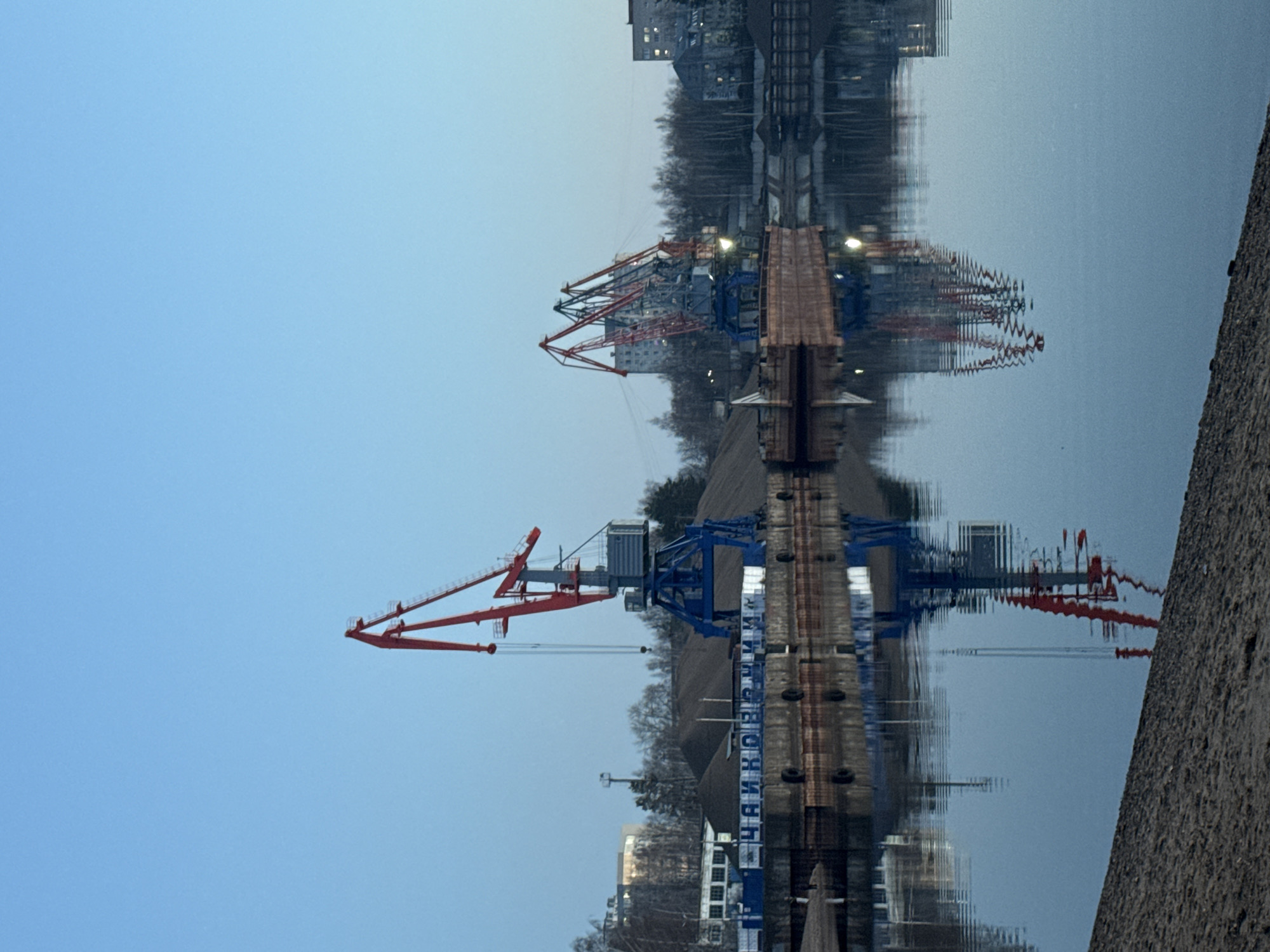 The image captures an industrial waterfront scene in Chaikovsky, Russia, under soft, ambient lighting suggestive of dusk or an overcast day. The scene features large industrial cranes and buildings reflected in still water.
A broad expanse of light blue sky fills the upper left portion of the frame. Along the main shoreline, a prominent red and blue gantry crane stands, its structure intricately detailed against the lighter sky. Further to the right and slightly behind, another large red crane is visible. The shoreline itself is lined with trees and various buildings, some displaying illuminated windows, hinting at a bustling city or town beyond the industrial infrastructure.
The lower right part of the image is dominated by the calm surface of the water, which creates a near-perfect, slightly distorted reflection of the entire scene above. The reflections of the cranes, buildings, and trees are clearly visible, with minor ripples adding to their texture.
Along the water's edge, a substantial concrete structure or pier runs horizontally. On this structure, the word "ЧАЙКОВСКИЙ" (Chaikovsky) is clearly inscribed in large Cyrillic letters, directly referencing the location context. To the far right, a textured concrete embankment or wall forms a sharp diagonal edge, bordering the water.
No people are visible, and no specific activity is taking place, giving the scene a quiet and contemplative atmosphere. The crisp reflections and the industrial elements provide the primary visual interest, showcasing a blend of functional architecture and tranquil natural surroundings.