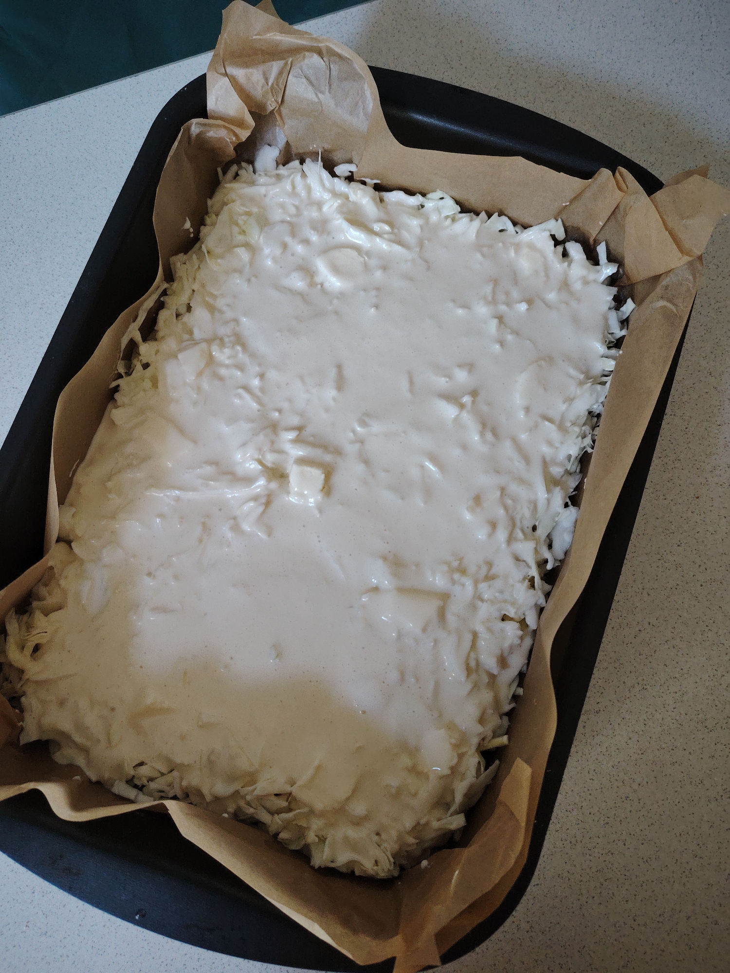 A high-angle shot shows a black baking pan lined with brown parchment paper, containing a rectangular dish. The dish is filled with a shredded white ingredient, possibly cabbage or potatoes, topped with a creamy, white batter or sauce. Small cubes of what appear to be cheese are visible within the topping. The baking pan rests on a speckled, beige countertop. The background shows a sliver of a dark green surface. There are no people or animals visible in the image. The scene is indoors, likely in a kitchen. The location in the city of Vladimirovka, Russia is not discernible from the image.