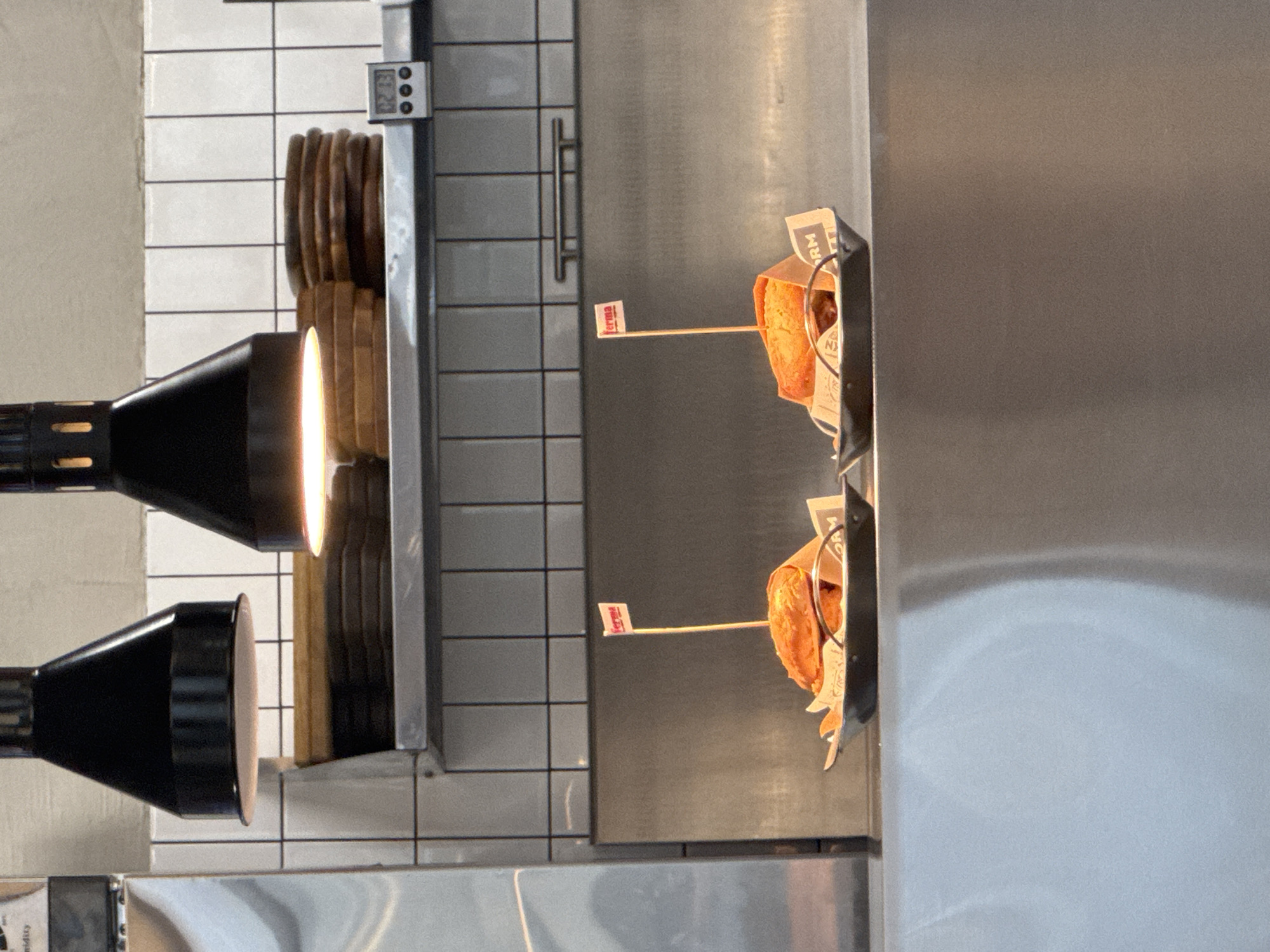 The image shows a close-up view of a food counter, likely in a restaurant. Two sandwiches, each with a small red and white flag bearing the word "ferma" stuck into them, are presented in black trays lined with brown paper. The sandwiches appear to be toasted. Behind the sandwiches, a stainless steel surface is visible, and above that, white tiled walls. Two large black lamps with conical shades hang overhead. On a shelf above the counter, a stack of round wooden objects and a digital timer are present. The overall scene suggests a food preparation or serving area. The location is identified as Kokshetau, Kazakhstan.