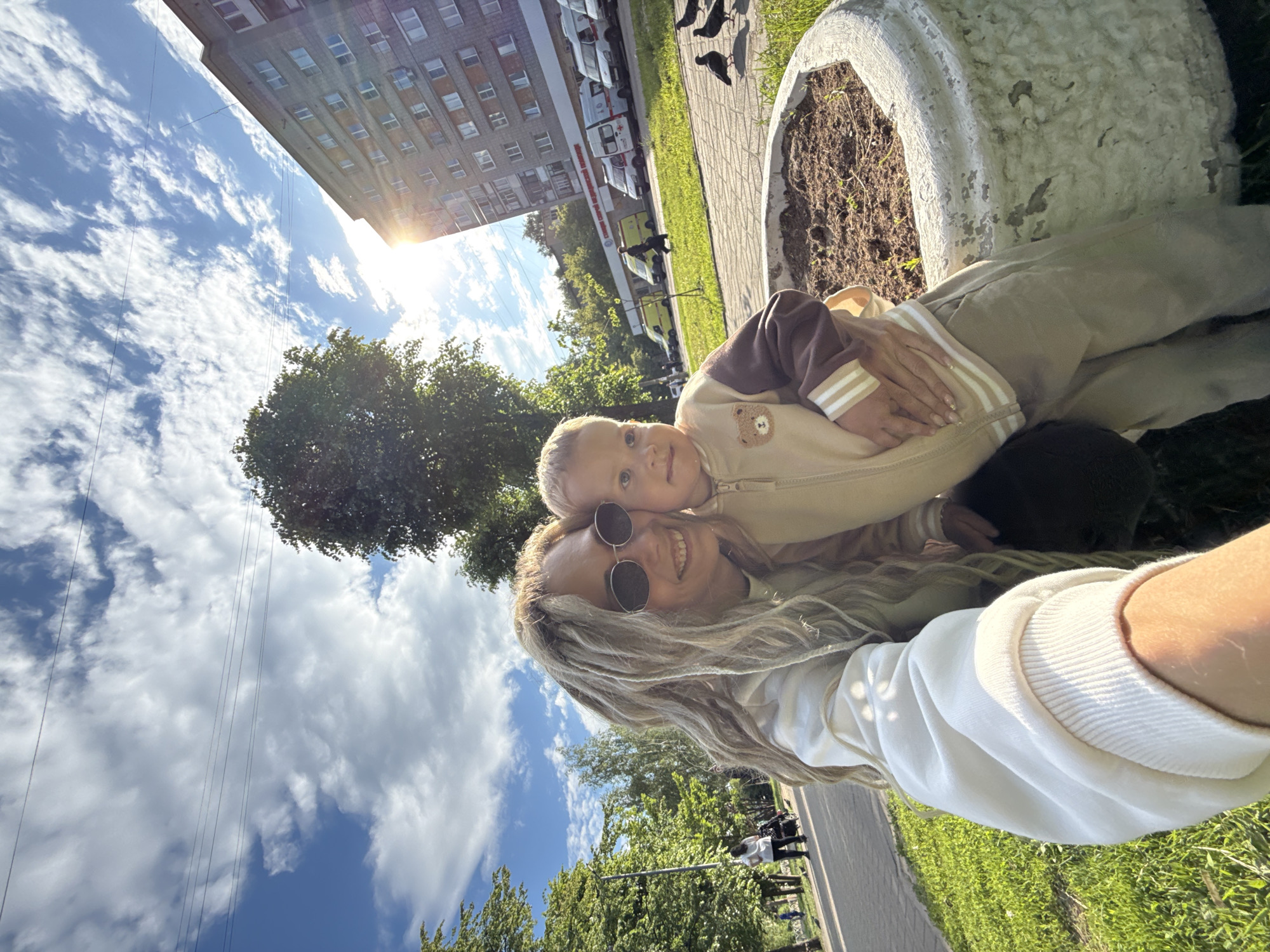 A brightly lit outdoor selfie captures a woman and a young child in what appears to be Rusanovo, Russia, on a sunny day. The image is oriented vertically, showing a blue sky with scattered white clouds and the sun's glare at the upper right.
The primary subjects are a woman and a child, positioned in the foreground. The woman, likely taking the photo, has long, light-colored hair, possibly dreadlocks or thick braids, and wears round sunglasses. She smiles broadly towards the camera, conveying happiness. Her left arm, clothed in a white long-sleeved top with a ribbed cuff, is visible. Her fingernails have a light manicure with subtle embellishments. The child, likely a boy with blonde hair, is nestled against her, embracing her with his left arm around her neck. He is wearing a light brown and beige tracksuit featuring a brown bear appliqué on the chest and white stripes on the sleeves. He looks directly at the camera with wide, inquisitive eyes and a faint smile.
The setting includes a paved path and green grass, suggesting a park or a residential area. To the subjects' right, a large, weathered concrete planter filled with soil is visible. In the background, beyond the path, several vehicles are parked along a street, including multiple white vans that resemble ambulances, some bearing red text on their side, partially visible as "МЕДИЦИНСКАЯ" (Medical). A multi-story residential building stands behind the vehicles. Several dark birds, likely pigeons, are seen on the paved path. Power lines crisscross the sky. Lush green trees are visible throughout the background and midground. The bright sunlight and clear sky indicate pleasant weather, possibly late morning or afternoon. The overall mood is joyful and affectionate.