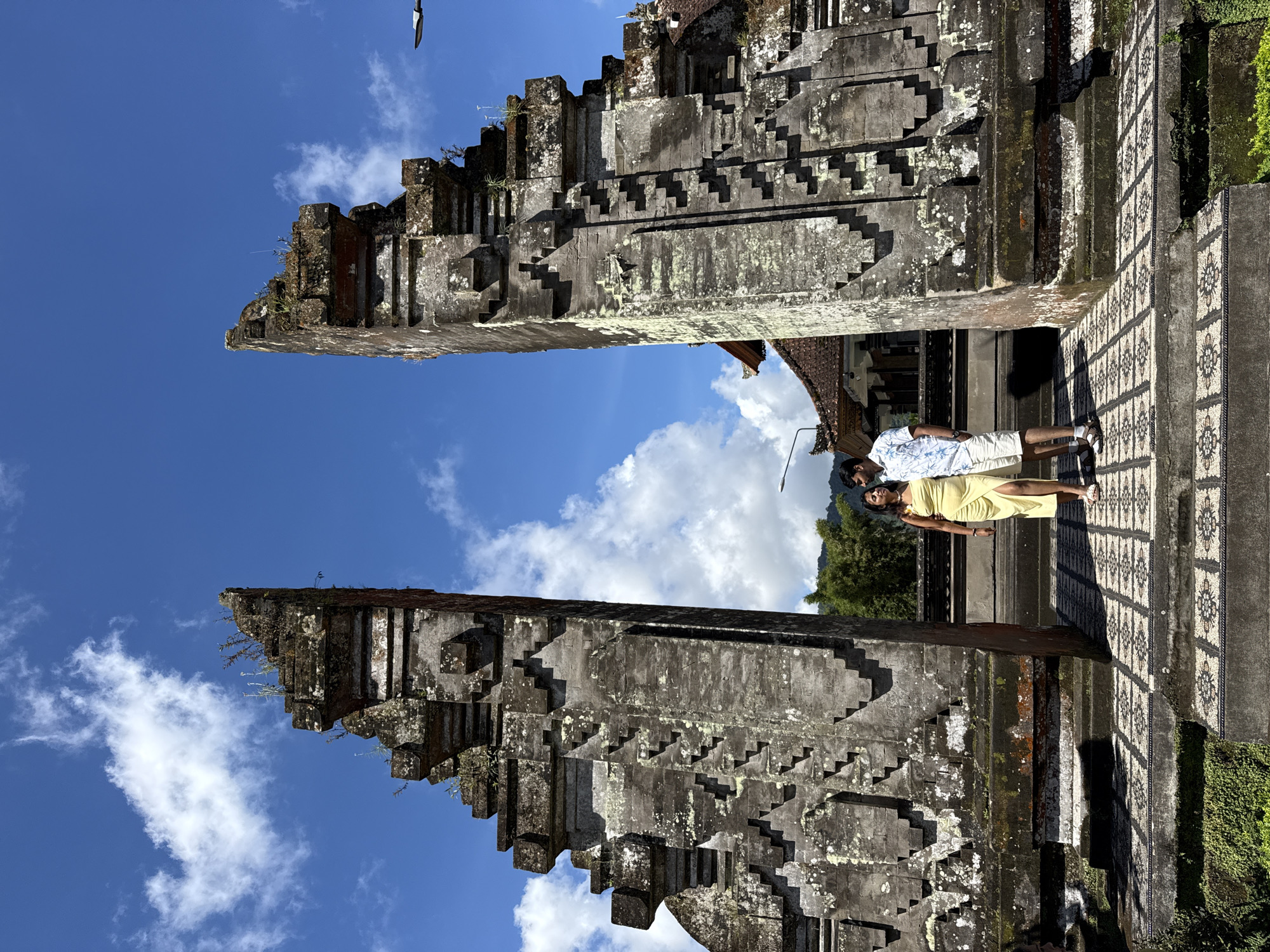 The image depicts a man and woman standing in the middle of the Handara Gate, located in Baturiti, Bali, Indonesia. The Handara Gate is a large traditional Balinese gate with a gray stone facade, decorated with a stepped pattern. The couple is dressed in vacation clothing and is standing on a decorative tile surface. The sky is a clear blue with white clouds visible through the gate opening.