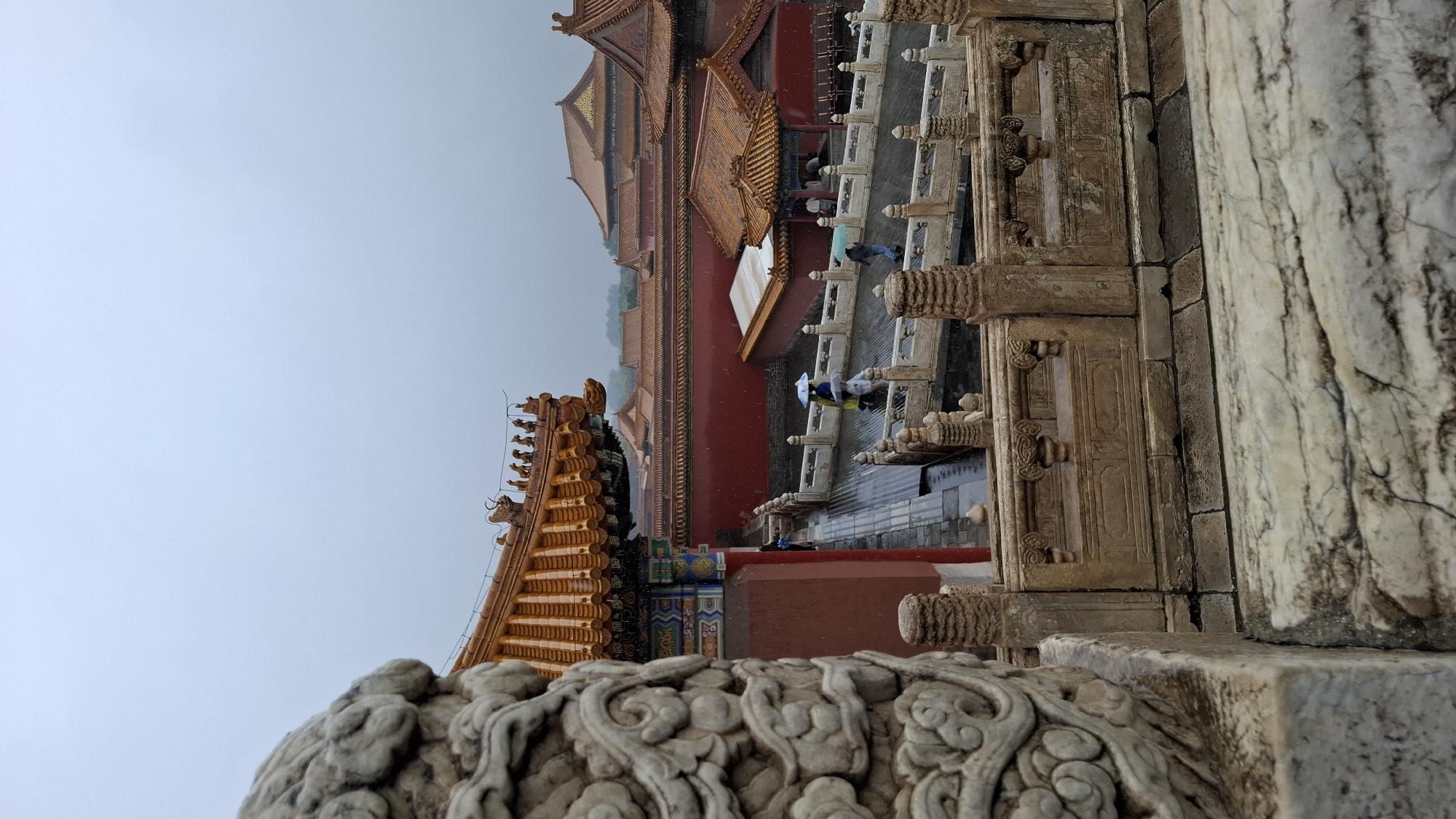 This image captures a rainy day at the Forbidden City in Beijing, Dongcheng District, China. The shot is taken from a low angle, emphasizing the intricate architectural details of the palace complex. In the foreground, a richly carved white stone balustrade and detailed doorway with decorative panels are visible. Beyond, the red walls and golden roofs of the Forbidden City buildings rise into the foggy sky. People with umbrellas can be seen walking along the elevated walkways.