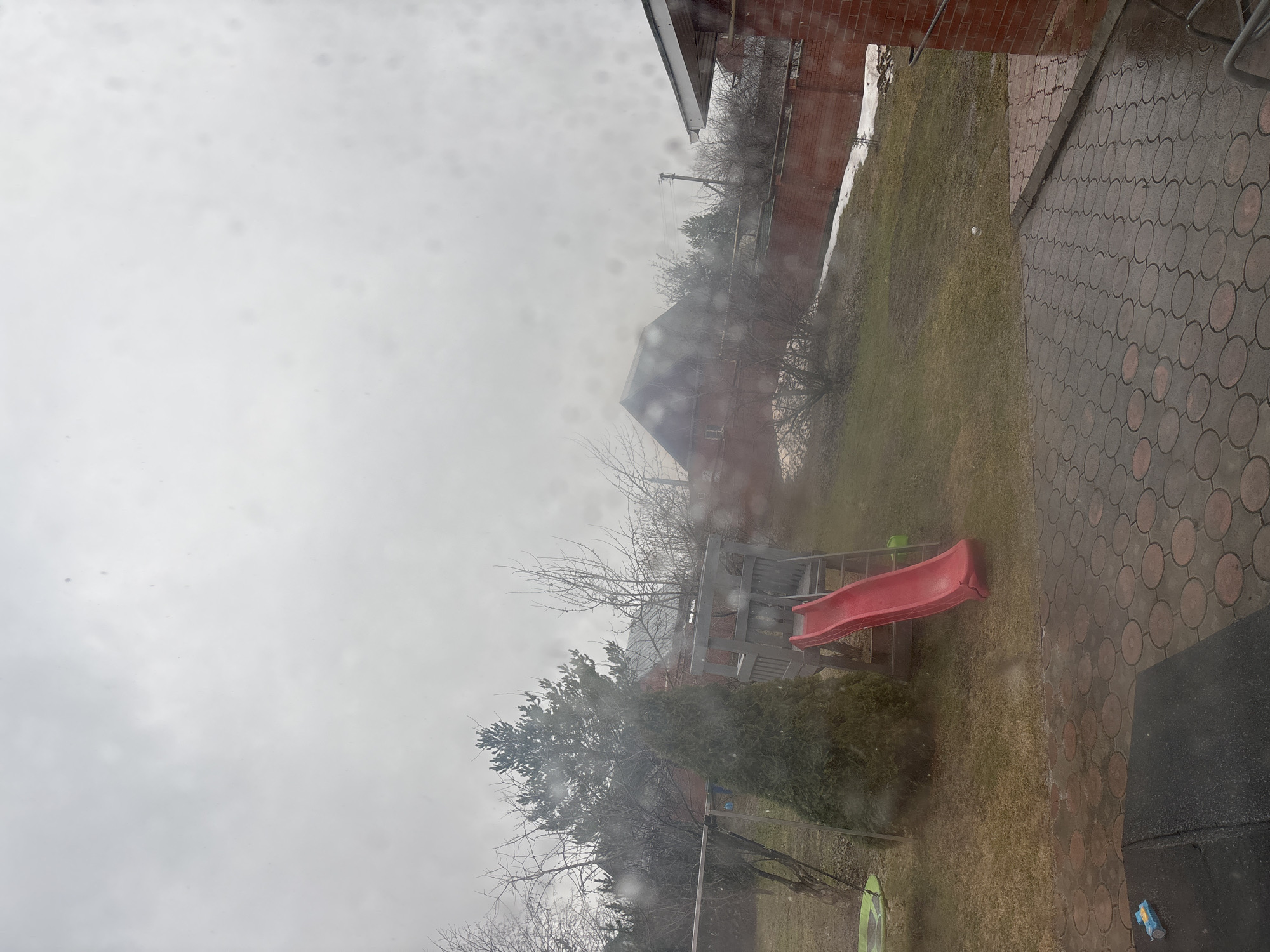 The image shows an overcast view from a window, likely in a residential area of Ferma, Russia, as specified in the prompt. The view includes a backyard with a grassy area, a red slide attached to a wooden play structure, and several trees and bushes, some bare of leaves. There is a paved area with a circular pattern to the right of the grassy area. Beyond the yard, there are other buildings visible, including a brick structure and a building with a pitched roof. The scene is likely being observed through a window with raindrops or condensation on it.