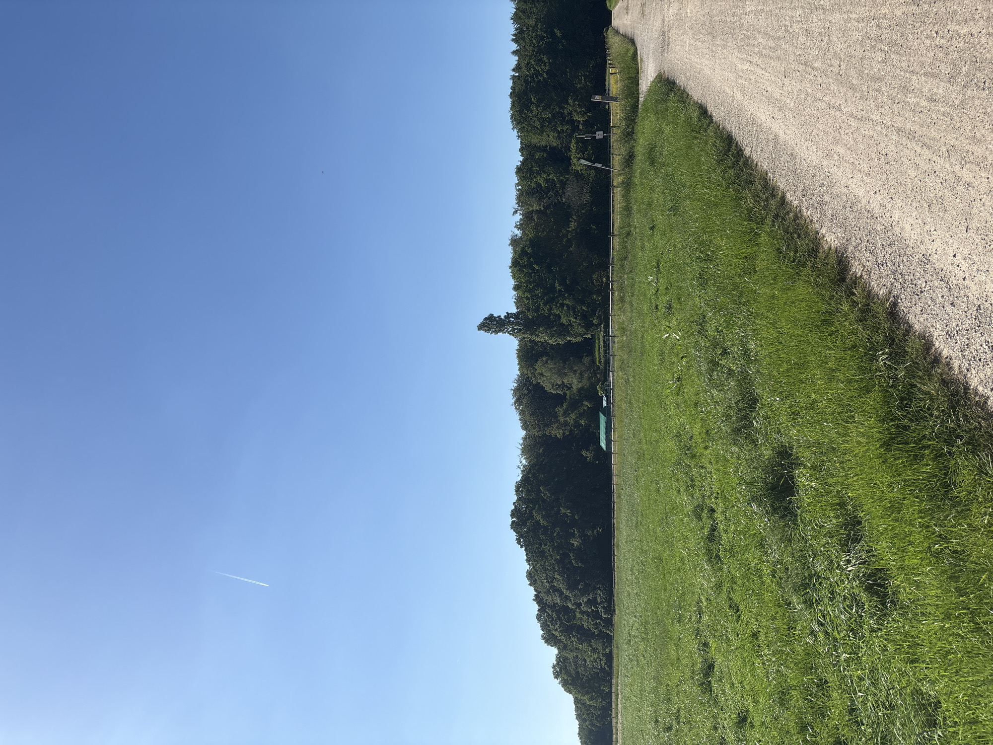The image shows a landscape with a bright blue sky, a field of green grass, a line of trees, and a gravel path. An airplane contrail is visible in the sky. Based on the environment, the location is likely in the city of Viersen, Germany. The scene depicts a rural or park-like setting. No people or interactions are directly visible in the image.