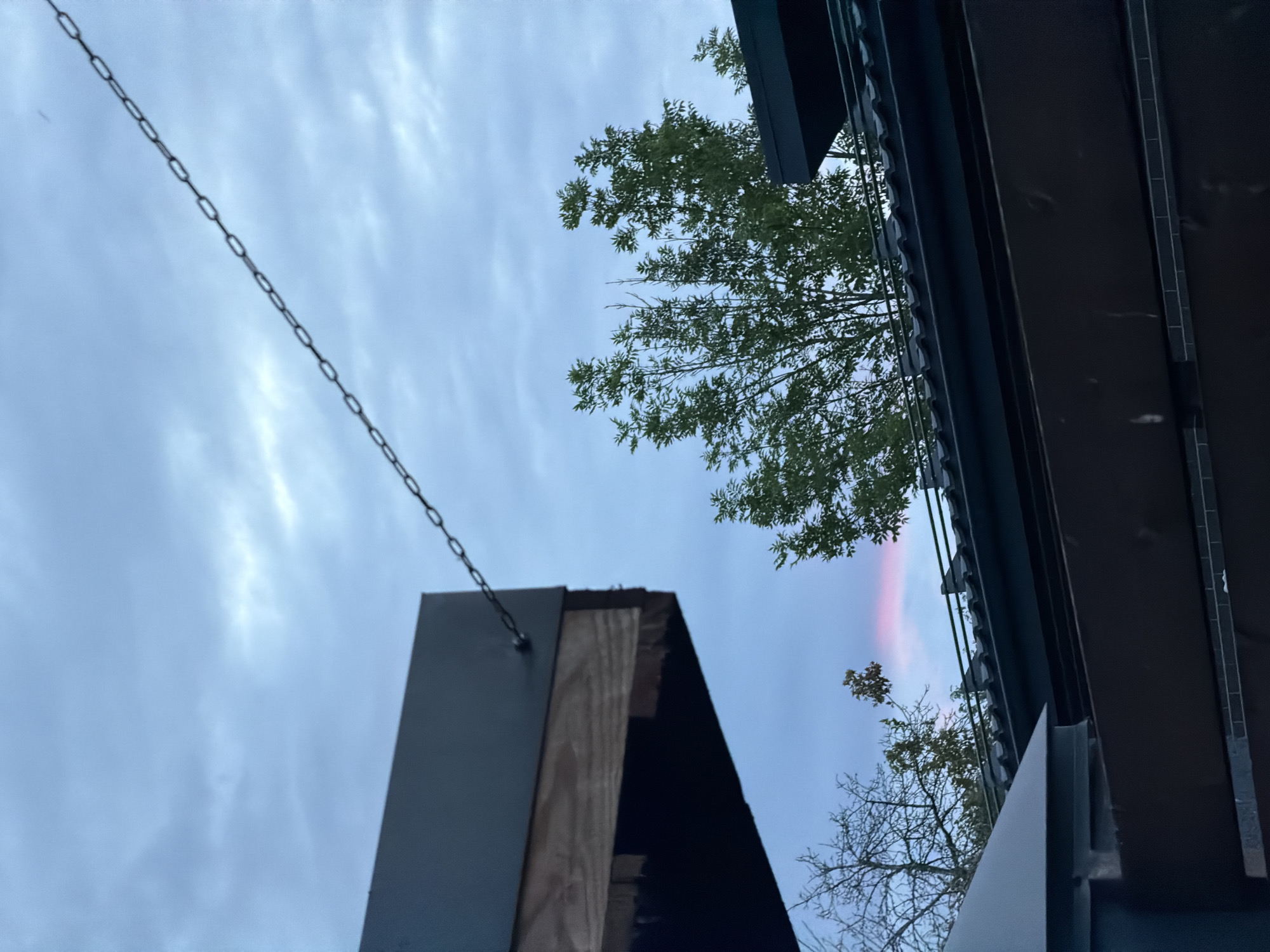 The image depicts an outdoor scene with a view of the sky and some man-made structures, likely in an urban or suburban setting. In the upper left quadrant, a dark, angled overhang of a building is visible, with visible wooden beams underneath and a metal chain extending diagonally upwards and to the right across the frame. The sky is overcast with pale blue and gray clouds, transitioning to a soft pink hue near the horizon in the lower center of the image, suggesting either dawn or dusk. Several trees with green foliage are interspersed throughout the scene, with some bare branches of a deciduous tree also visible on the left. Below the trees, dark-tiled roofs of buildings are present. In the foreground, dark brown horizontal elements, possibly a fence or a deck railing, are prominent, with a lighter colored surface above it. There are no people or animals visible in the image. The overall impression is a still, twilight or dawn scene focusing on architectural elements and the sky.