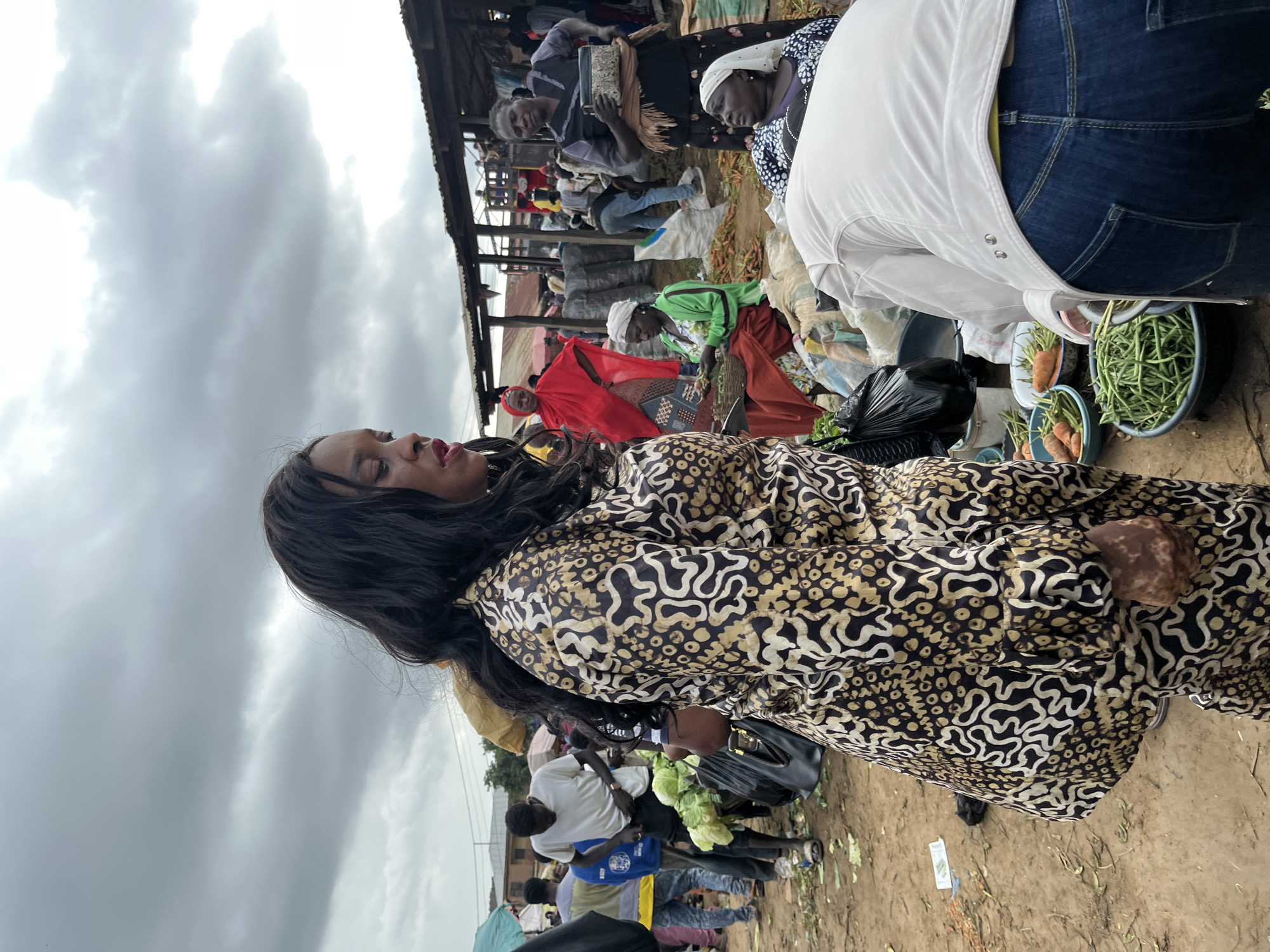 The image depicts a bustling outdoor market scene, likely in Gura Top, Nigeria, under a cloudy sky suggesting overcast or impending rain. The foreground features a woman with long, dark hair, wearing a distinctive black and gold patterned dress. She is looking to her left, with a neutral expression. To her right, and further into the background, other people are visible, engaged in various market activities.
Several individuals are dressed in traditional attire, including women wearing headscarves and long dresses. One woman in a bright red garment stands out. There are also men and women in more casual clothing, such as jeans and t-shirts. Objects in the scene include baskets of produce, notably carrots and green beans, suggesting the sale of fresh vegetables. A black garbage bag is also visible, indicating waste management within the market. The setting is a covered market area, with wooden structures and stalls creating an organized yet busy atmosphere.
The overall activity appears to be daily commerce, with vendors and shoppers interacting. The image captures the vibrant energy of a local marketplace. There are no discernible signs with visible text. The time of day is not explicitly indicated, but the cloudy weather suggests it could be morning or afternoon. The scene is rich with the textures of fabrics, produce, and the earthy ground.
