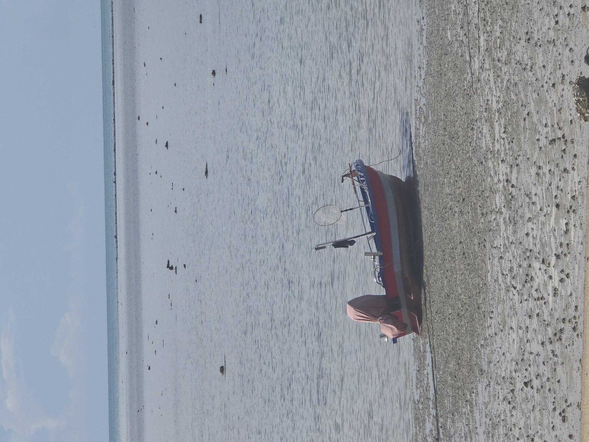 A single, red and white motorboat is moored near the shore at low tide in Baan Chaweng Noi, Thailand. The boat, partially covered with a pink tarp, sits on a muddy, pebble-strewn shoreline. A fishing net is attached to a pole extending from the boat. The calm, shallow water stretches to a distant horizon under a pale blue sky. Scattered rocks are visible in the water and on the shore.