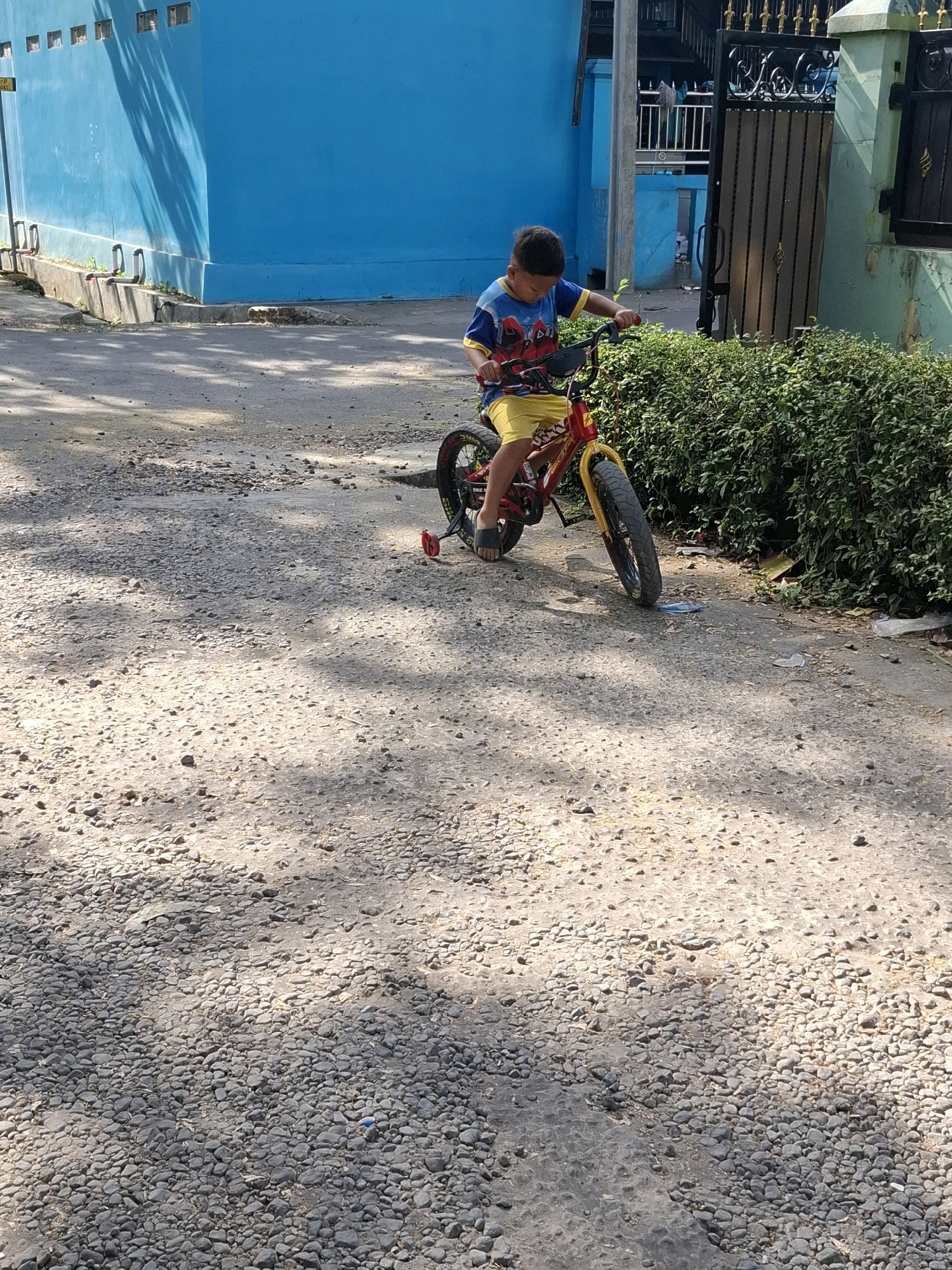 A young boy wearing a blue and yellow t-shirt with a superhero print and yellow shorts is riding a red and yellow bicycle with training wheels on a gravel road in front of a bright blue wall. The boy is looking down at the handlebars. To the right of the boy is a dense green hedge. In the background, there is a black metal gate with ornate detailing and a green wall. The scene is outdoors, with shadows cast on the road from unseen trees. The overall impression is a casual outdoor scene, possibly in a residential area of Karangtengah, Indonesia.