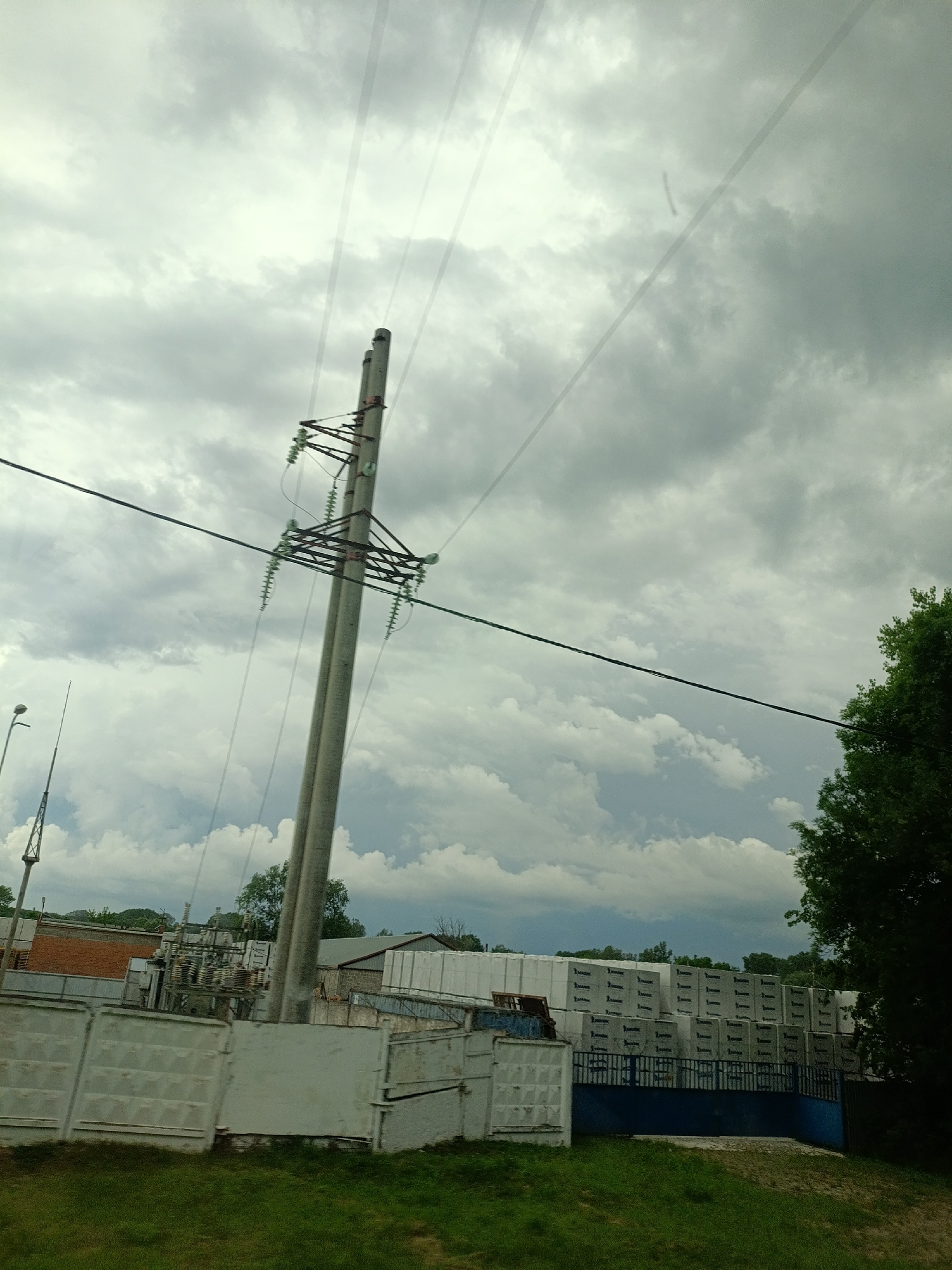 A medium-shot, eye-level view captures an industrial area on a cloudy day. Dominating the foreground is a double-poled electrical pylon, its concrete structure supporting a complex network of wires and insulators. To the left, a street lamp with a tall, slender pole stands in front of a smaller, lattice-style tower. Beyond the pylon, a white concrete fence with embossed patterns runs horizontally across the frame. Behind the fence lies an industrial complex featuring brick buildings and a structure with stacked, white, rectangular modules, each marked with black lettering. A dark blue metal fence encloses a portion of the complex, and a vibrant green tree occupies the right edge of the frame, partially obscuring a view of the sky. The sky itself is filled with a thick layer of grey clouds. The scene appears to be located in Saratovskaya, Russia.
