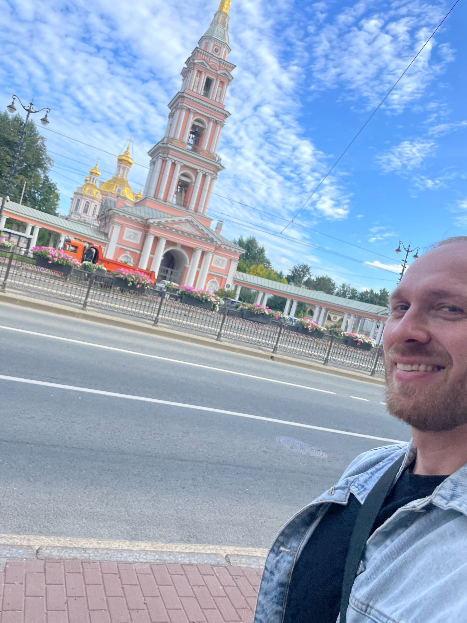 In a selfie taken from a low angle, a man with a beard and a light blue denim jacket smiles at the camera. He is positioned in the lower right corner of the frame, partially obscuring the background. Behind him, a multi-story building with pink and white accents, topped with golden domes, is visible. A tall, slender tower, also with pink and white detailing, rises prominently in the background, featuring arched windows and a golden spire. The building appears to be a church or cathedral. A street with traffic is in the mid-ground, bordered by a black metal fence adorned with planters filled with pink and white flowers. An orange truck is parked on the side of the road. The sky above is bright blue with scattered white, wispy clouds. The scene is set on a paved sidewalk, with the man standing near a red brick walkway. The overall impression is of a sunny day in an urban environment, likely Saint Petersburg, Russia, given the architectural style.
