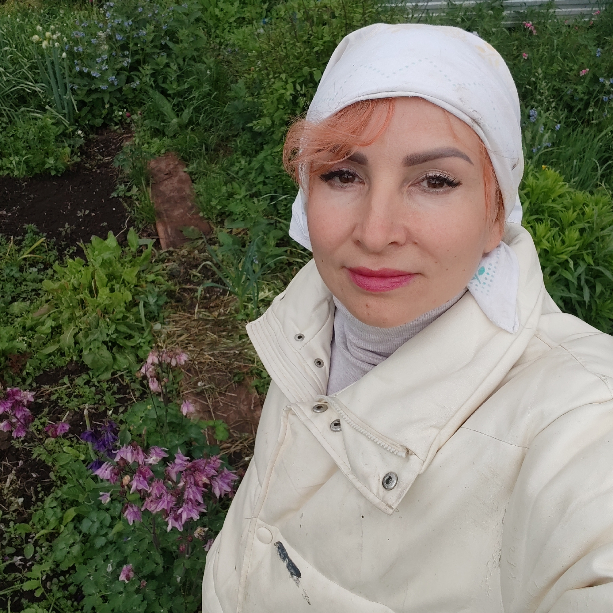 This image captures a woman in a garden located in the city of Chaikovsky, Russia. The woman, positioned centrally in a close-up, is facing the camera. She has fair skin, reddish-orange hair visible on her forehead, and dark eyes with noticeable eyeliner and possibly false eyelashes. Her lips are painted a bright pink. She wears a white headscarf with a subtle pattern, a light grey turtleneck, and an off-white quilted jacket secured by snap buttons and a zipper. The jacket shows signs of wear, including a dark smudge on its lower left side. The background consists of a garden with dark soil. Various green plants are present, including tall green stalks resembling onions on the left, numerous leafy greens, and clusters of blue forget-me-nots in the mid-background. Purple and pink columbine flowers are visible in the lower left and lower right foreground. A piece of reddish-brown wood lies on the ground in the mid-right. A light-colored fence or structure is partially discernible in the upper background.