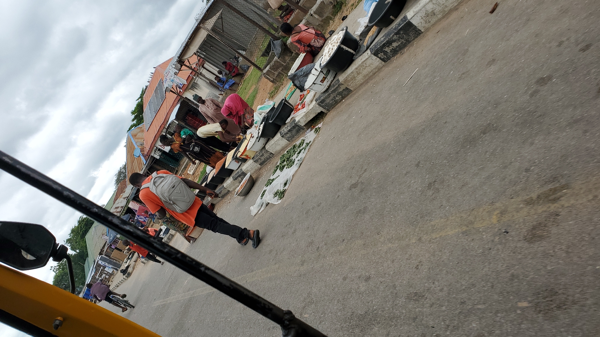 This image, taken in Jalingo, Nigeria, depicts a street market scene on an overcast day. In the foreground, a person wearing an orange shirt and a backpack is walking away from the camera on the asphalt road. To the right, a curb separates the road from a market area where several people are engaged in commerce. Various goods, including vegetables and grains, are displayed in plastic basins and on white sheeting on the ground. Buildings with corrugated iron roofs line the background, some appearing to have solar panels. To the left, a person on a bicycle is riding down the street. The overall atmosphere suggests a typical bustling marketplace.