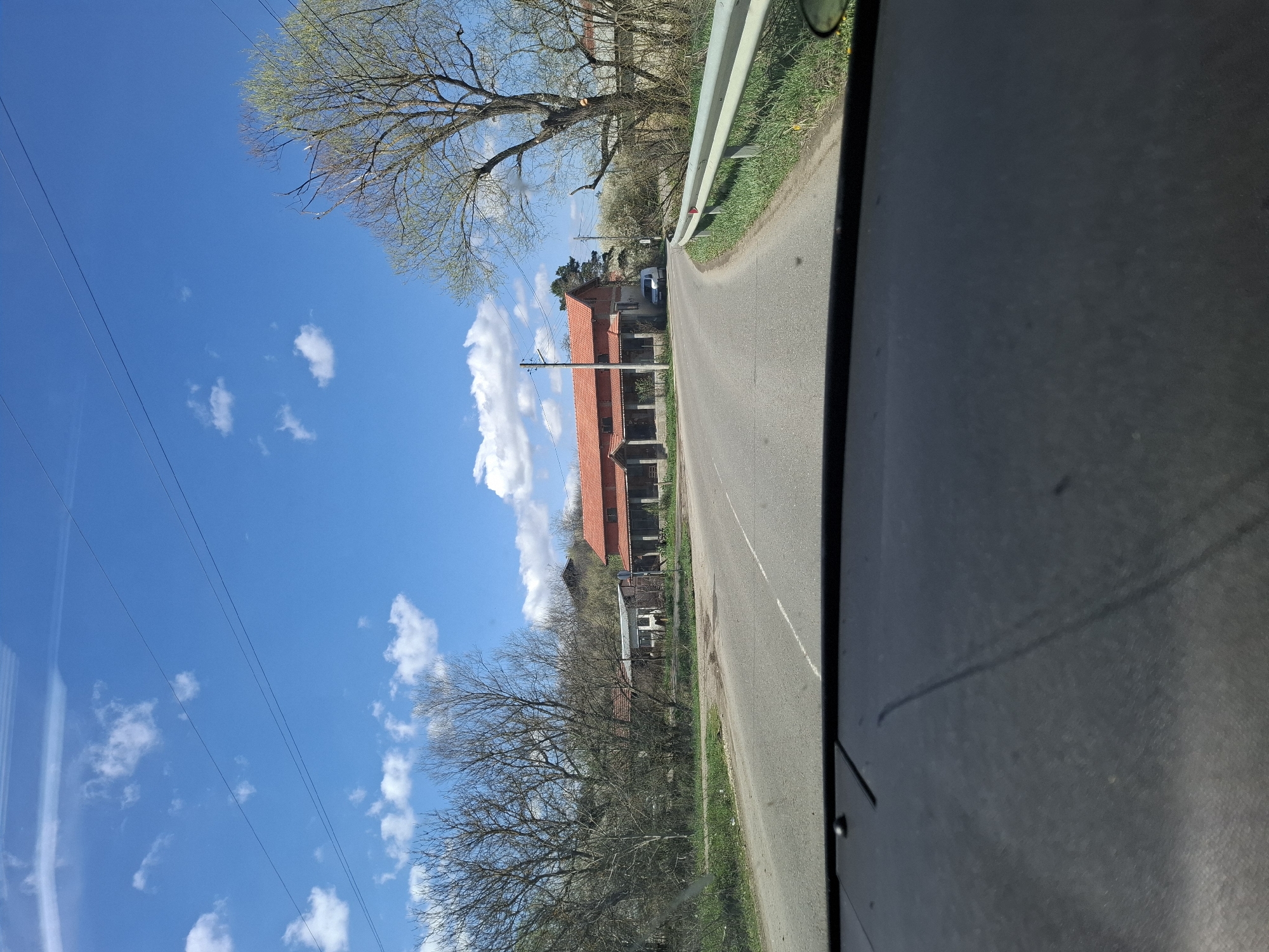 The image presents a view from inside a vehicle traveling on a rural road near Studeno Buche, Bulgaria. On the left, a clear blue sky with scattered white clouds is visible, punctuated by utility lines. Trees with bare branches line the roadside, interspersed with buildings having red-tiled roofs. The asphalt road stretches ahead, bordered by a metal guardrail on the right. The interior of the vehicle, including a portion of the dashboard or door panel, occupies the right side of the frame. A single vehicle is visible ahead.