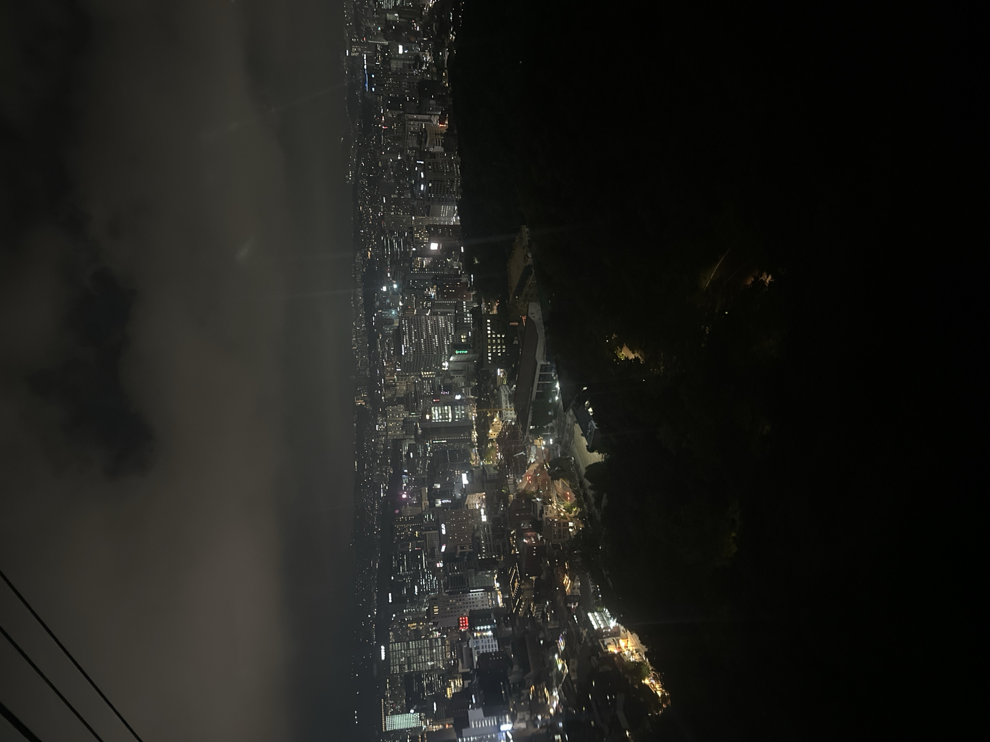 The image shows an aerial night view of Seoul, South Korea. The city is illuminated with bright streetlights, and the skyline is partially obscured by clouds or fog on the left side of the image. There is a large, dark green area, possibly a forest, on the right side. In the bottom left corner, there are black diagonal lines, which appear to be cables or wires, likely from a cable car or aerial tram. The scene suggests an observation point, perhaps a mountain or tower, overlooking the city at night.