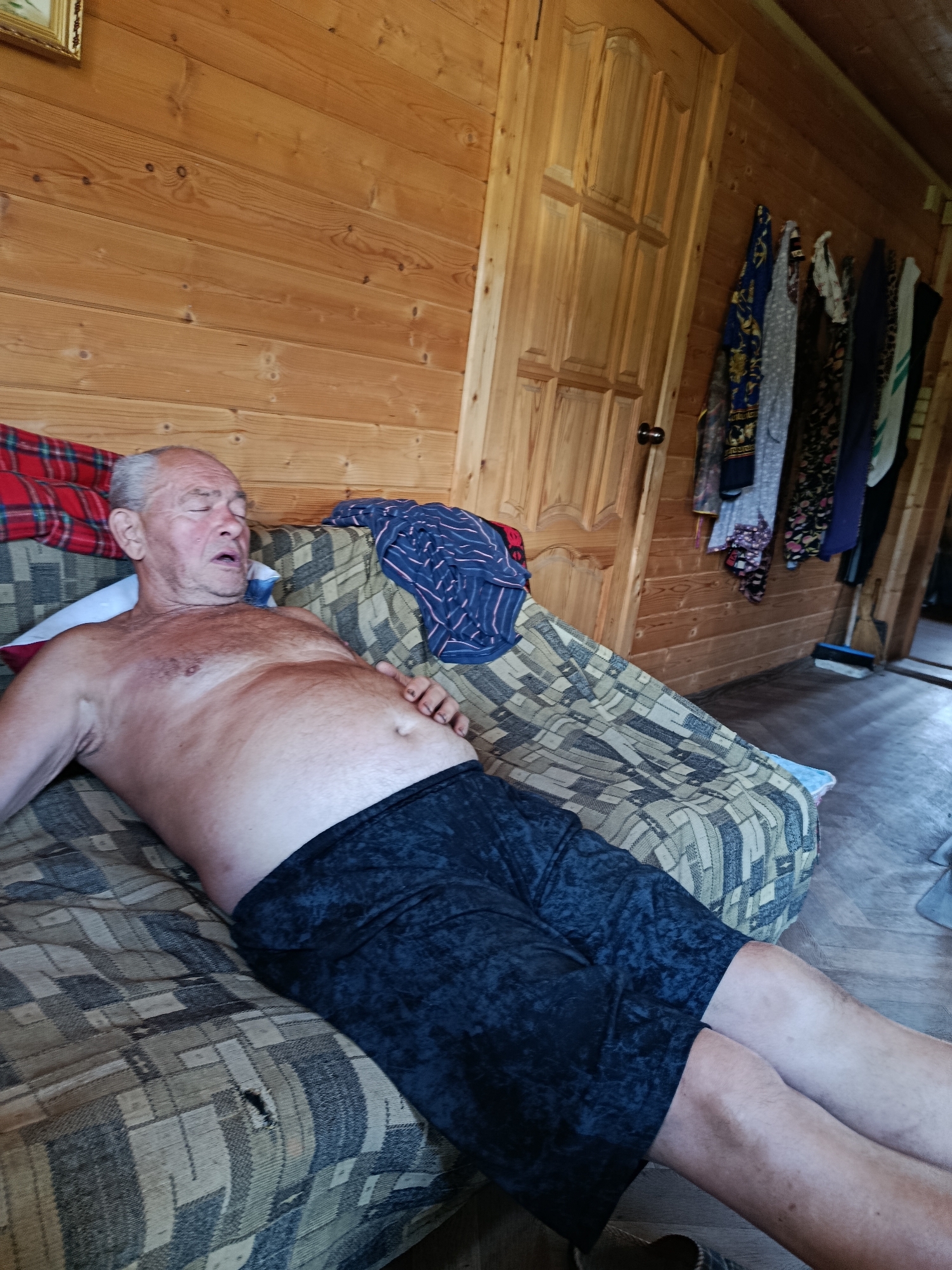 An elderly man with gray hair, bare-chested and wearing dark shorts, is asleep on a couch in a room with wood-paneled walls. He is lying on his back with his eyes closed and mouth slightly open. A pillow is behind his head, and a patterned blanket covers the couch. To the right of the man, a wooden door is visible, and several items of clothing hang on hooks on the wall. A broom and dustpan are propped up in the corner of the room. The scene appears to be in a rustic dwelling, possibly a cabin or a country house.