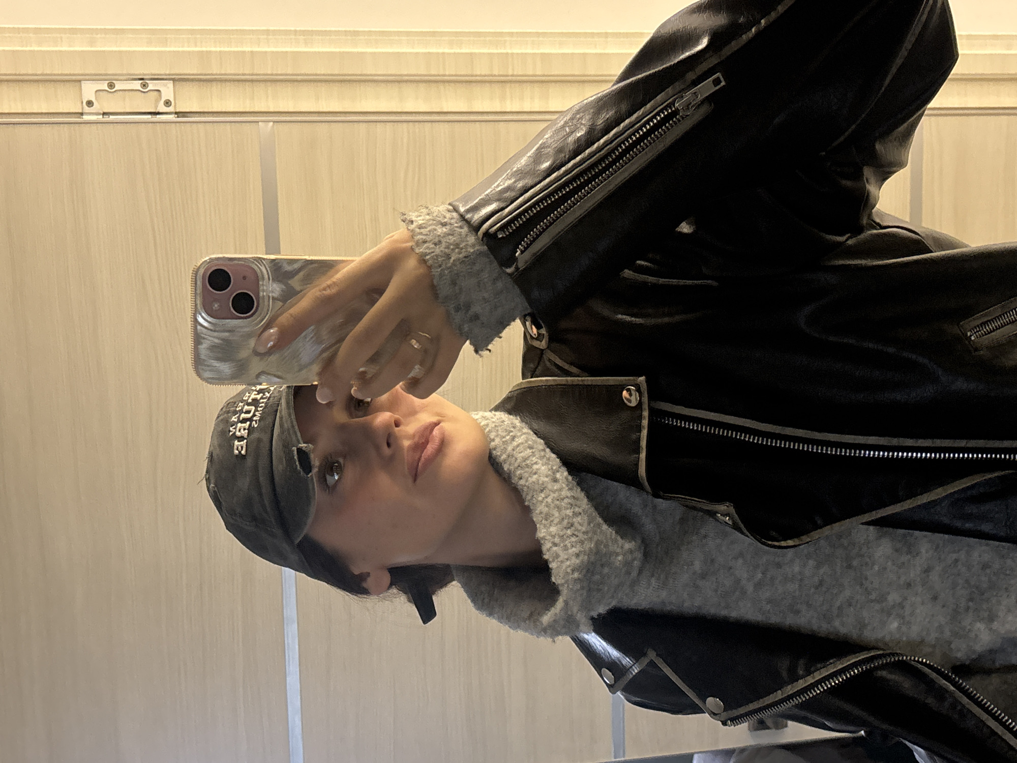 A woman is taking a selfie with a smartphone in front of a light-colored wall. She is wearing a black leather jacket over a gray sweater and a gray baseball cap. The smartphone is silver. The location is not identifiable as Krasnoyarsk, Russia based on the visual information provided.