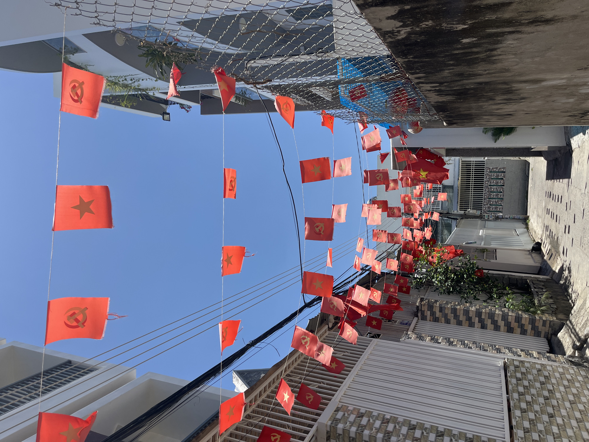 The scene is an outdoor street view in Bac Nha Trang Ward, Vietnam. Red flags are strung across the street from above. The flags contain both a yellow star and a yellow hammer and sickle symbol. Buildings line the street on either side. The sky is a clear blue.