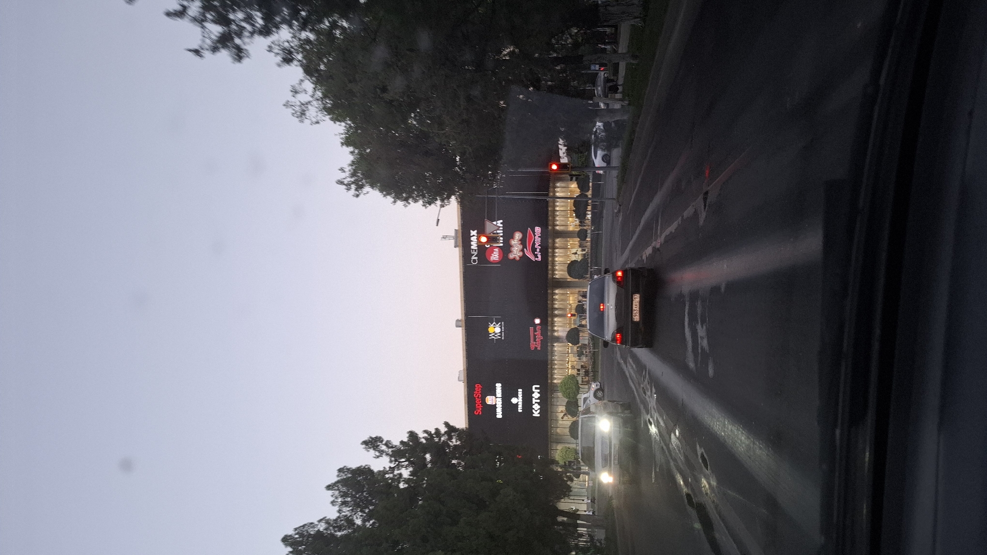 The image depicts a street scene in the city of Shymkent, Kazakhstan, at dusk. The foreground is dominated by a car's dashboard and windshield, suggesting the photo was taken from inside a vehicle. The wet asphalt road shows reflections of lights from vehicles and streetlights. In the mid-ground, two cars are visible. The car in front has its taillights illuminated, and a license plate reading "963AKG 17" is visible. To the left, another car with its headlights on is moving forward.
Dominating the background is a large, modern building complex, identified as a shopping center. It features illuminated signage for various establishments, including "SuperStep," "BURGER KING," "STARBUCKS," "KOTON," "WOK WOK," "CINEMAX," "TAPA.TAPAS," "Lady Fragrance," and "LI-NING." A traffic light is visible on the right side of the image, displaying a red signal. Trees line the sides of the road, and the sky above is a soft, pale blue, indicative of twilight. The overall atmosphere is one of urban activity transitioning into the evening.