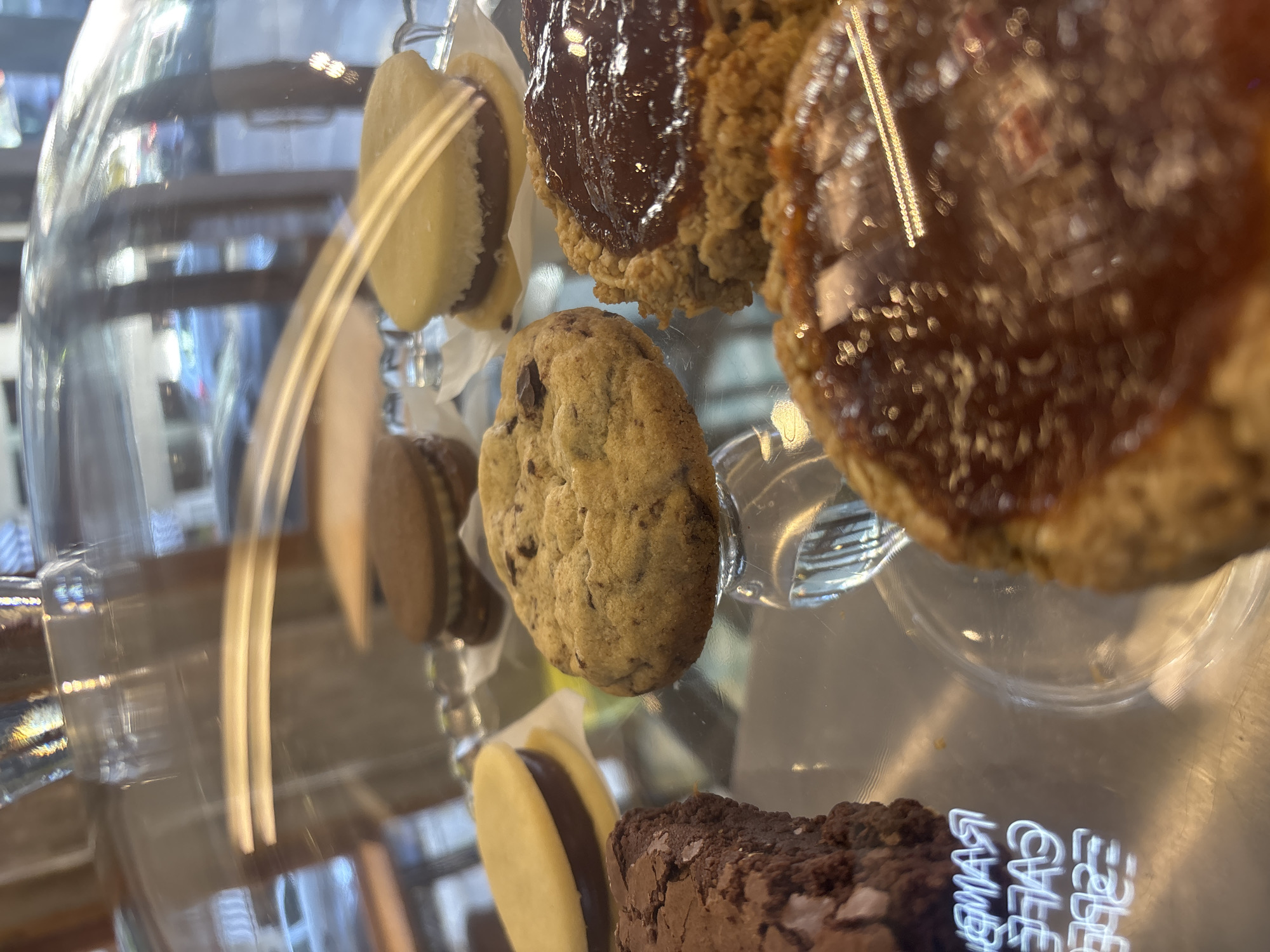 The image shows a display case of pastries. The pastries include sandwich cookies with a light yellow shell and dark filling, a chocolate chip cookie, jam-topped cookies, and a brownie. The text "MART CAFÉ" is visible on the bottom shelf of the display case. The pastries are likely in a cafe in Buenos Aires, Argentina.