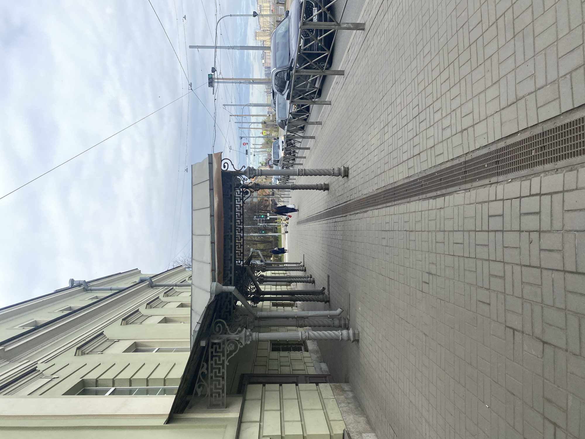 This image, taken in Saint Petersburg, Russia, depicts an urban street scene on an overcast day. The perspective is looking down a sidewalk parallel to a building on the left and a street on the right.
On the left, a light-colored (pale green or beige) building with multiple windows and prominent architectural cornices extends upwards. Below its roofline, an ornate metal canopy or awning stretches over the sidewalk, supported by a series of decorative pillars. These pillars alternate between fluted and twisted designs, adding a classical touch. The canopy's edge features a repeating Greek key pattern. A person in a dark coat is visible walking beneath this canopy in the mid-ground.
The sidewalk is paved with light grey interlocking pavers arranged in a herringbone pattern. A long metal drainage grate runs along the edge of the sidewalk, separating it from the street. To the right of the sidewalk, a row of dark metal barriers or fences delineates the street from the pedestrian area. Several cars are parked along the curb, and one car's license plate shows "321 198" along with the Russian flag and "RUS" letters.
Overhead, a complex network of wires, likely for trams or trolleybuses, crisscrosses the sky, supported by numerous utility poles. In the distance, several traffic lights are visible, some displaying a green signal. Other indistinct figures of pedestrians are seen further down the street. The sky is uniformly overcast with light grey clouds, indicating daytime but not direct sunlight. The scene captures a typical, unhurried moment of daily life in a city.