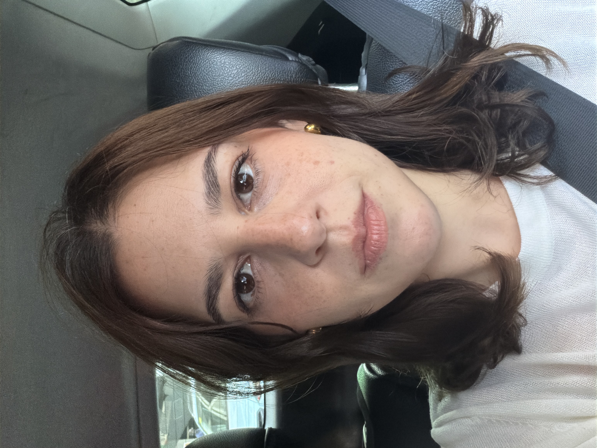 A selfie is taken from the perspective of someone sitting in the passenger seat of a car. The subject, a woman with brown hair and freckles, is facing the camera. She is wearing a white shirt and a gold hoop earring. The background shows the interior of the car, including a black leather headrest and a seatbelt. Outside the car, through the window, a blurred view of city traffic can be seen. The image does not contain enough information to determine the location within Barcelona, Spain.