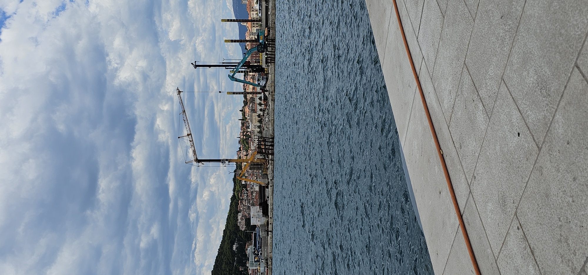 This is a vertical, eye-level, outdoor, daytime shot of a harbor in Split, Croatia. In the foreground, there is a light-colored, stone-tiled walkway that slopes gently upward from left to right. A thick, orange hose lies diagonally across the walkway. The middle ground is dominated by the dark blue, choppy water of the harbor. In the background, a construction site is visible on the shoreline, featuring several large cranes of varying types and colors, including a prominent red and black tower crane and a turquoise excavator. Piles of building materials and unfinished structures are also present. Beyond the construction, a densely built city with red-tiled roofs extends up a green, tree-covered hill. The sky is filled with dramatic, grey and white clouds, with patches of blue sky visible.