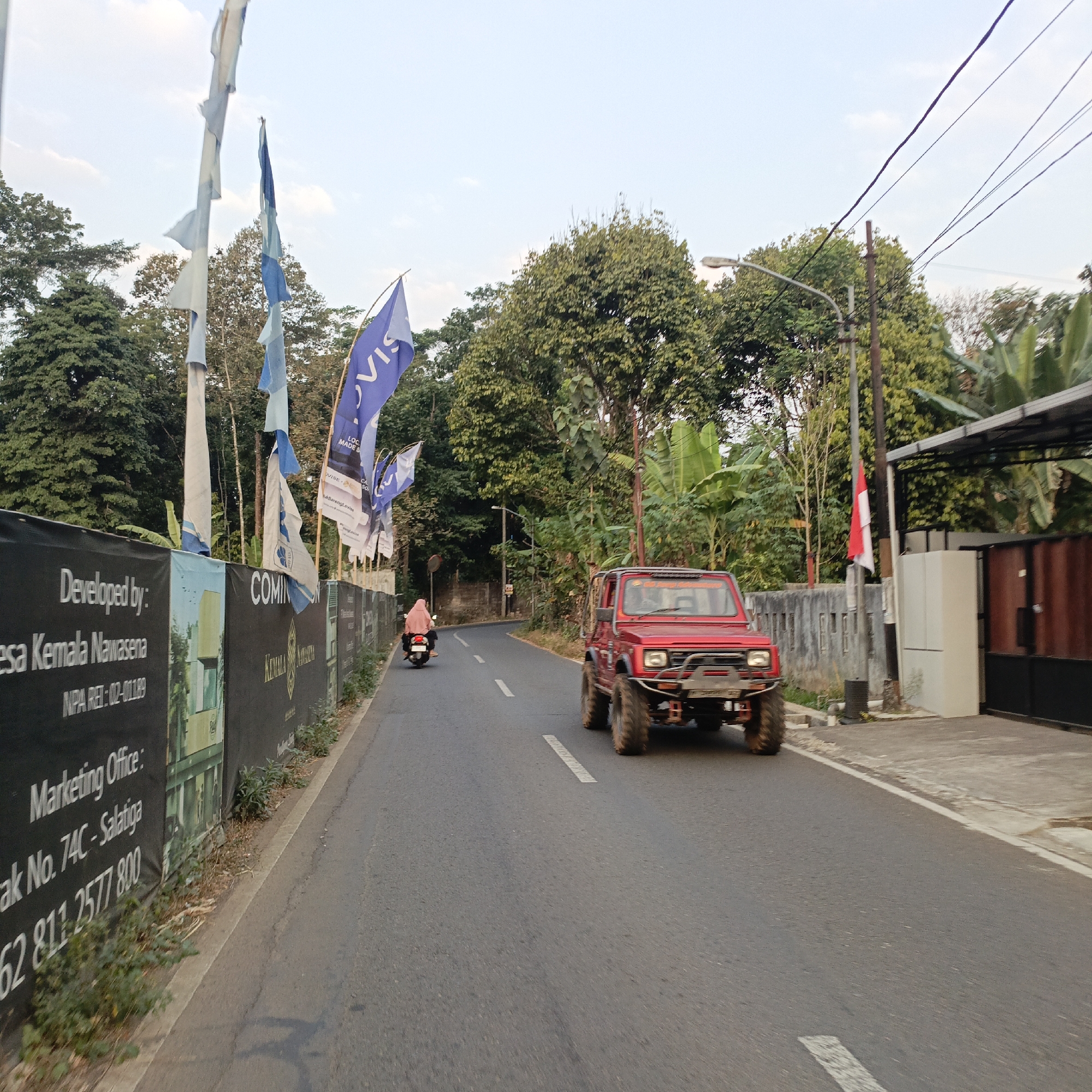 A red, modified off-road vehicle with large tires is driving on a paved road. To the left of the road, there is a black banner with white text that reads "Developed by: Kesa Kemala Nawasena", "NPA REI: 02-01189", and "Marketing Office: ...". Behind the banner are several flags, some of which are torn, and a line of trees. A person is riding a scooter on the left side of the road. To the right of the road, there is a grey fence, a red and white flag, a building with a dark roof, and more trees. The sky is overcast. The scene appears to be in an urban or suburban area, possibly a construction site, given the banner and flags. The text on the banner and the mention of "Salatiga" in the text indicates the location is Salatiga, Indonesia.