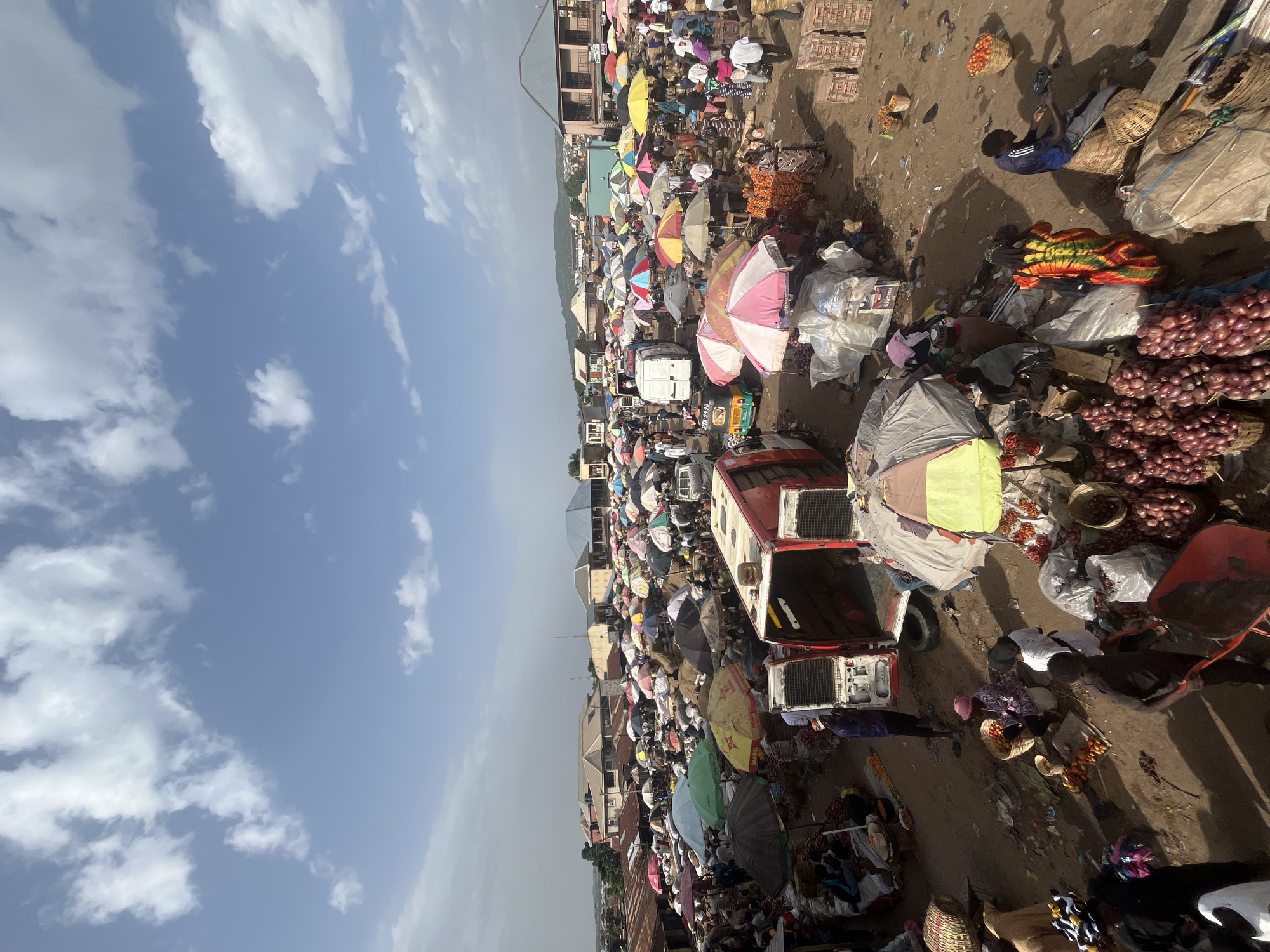 This is an aerial, wide-angle shot of a crowded outdoor market in Rafin Par, Nigeria. The scene is bustling with activity under a bright, partially cloudy sky. Numerous vendors are operating from stalls, many covered by colorful umbrellas providing shade. Various goods are on display, including large piles of red onions, baskets of tomatoes, and other produce. Several vehicles, including vans and a three-wheeled auto-rickshaw, are present within the market, some appearing to be used for transport or as mobile stalls. People are seen moving through the market, interacting with vendors, and carrying goods. The ground is dusty and appears to be a mix of dirt and sand, with some litter scattered about. In the background, buildings with corrugated metal roofs and some more permanent structures are visible, suggesting a densely populated urban or semi-urban environment. The overall impression is of a vibrant and active marketplace.