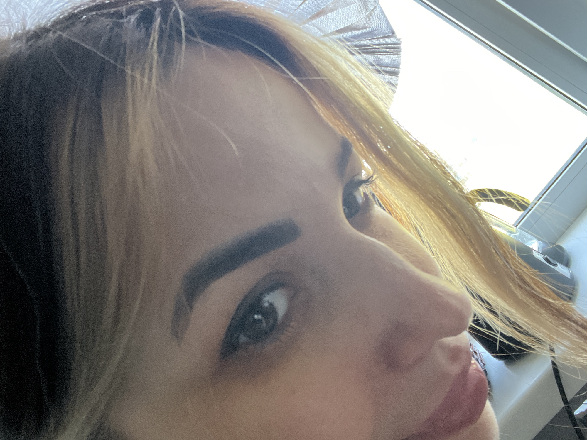 A close-up, angled selfie shows a person with fair skin and dark eyebrows. The individual has dark hair at the roots transitioning to blonde highlights. The eyes are dark and appear to be looking upwards. To the upper right of the frame, a bright window is visible, and an object resembling a hair dryer sits on a shelf in front of the window. The location is Unknown, Unknown.