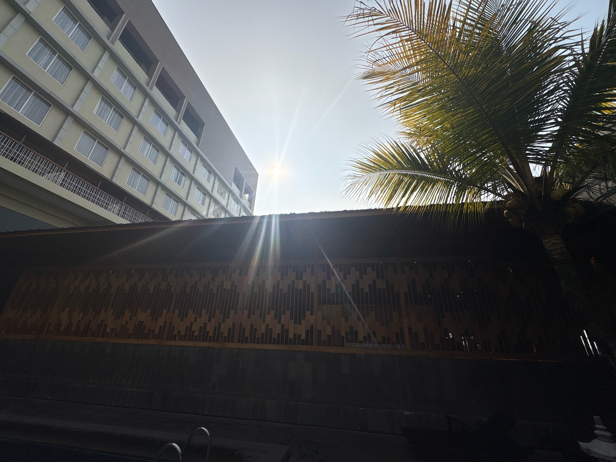 In Pamekaran, Indonesia, the image captures a low-angle view of a modern building with a facade of light-colored horizontal panels and numerous rectangular windows, each covered by white curtains. Above the building, a bright sun shines through a clear sky, creating lens flare effects. To the right, a palm tree with large fronds extends into the frame, its leaves illuminated by the sun. In the foreground, a dark, textured wall with a patterned bamboo-like screen runs horizontally. Below this wall, a dark swimming pool is visible with a silver pool ladder. The scene appears to be a resort or hotel setting, possibly during the daytime.