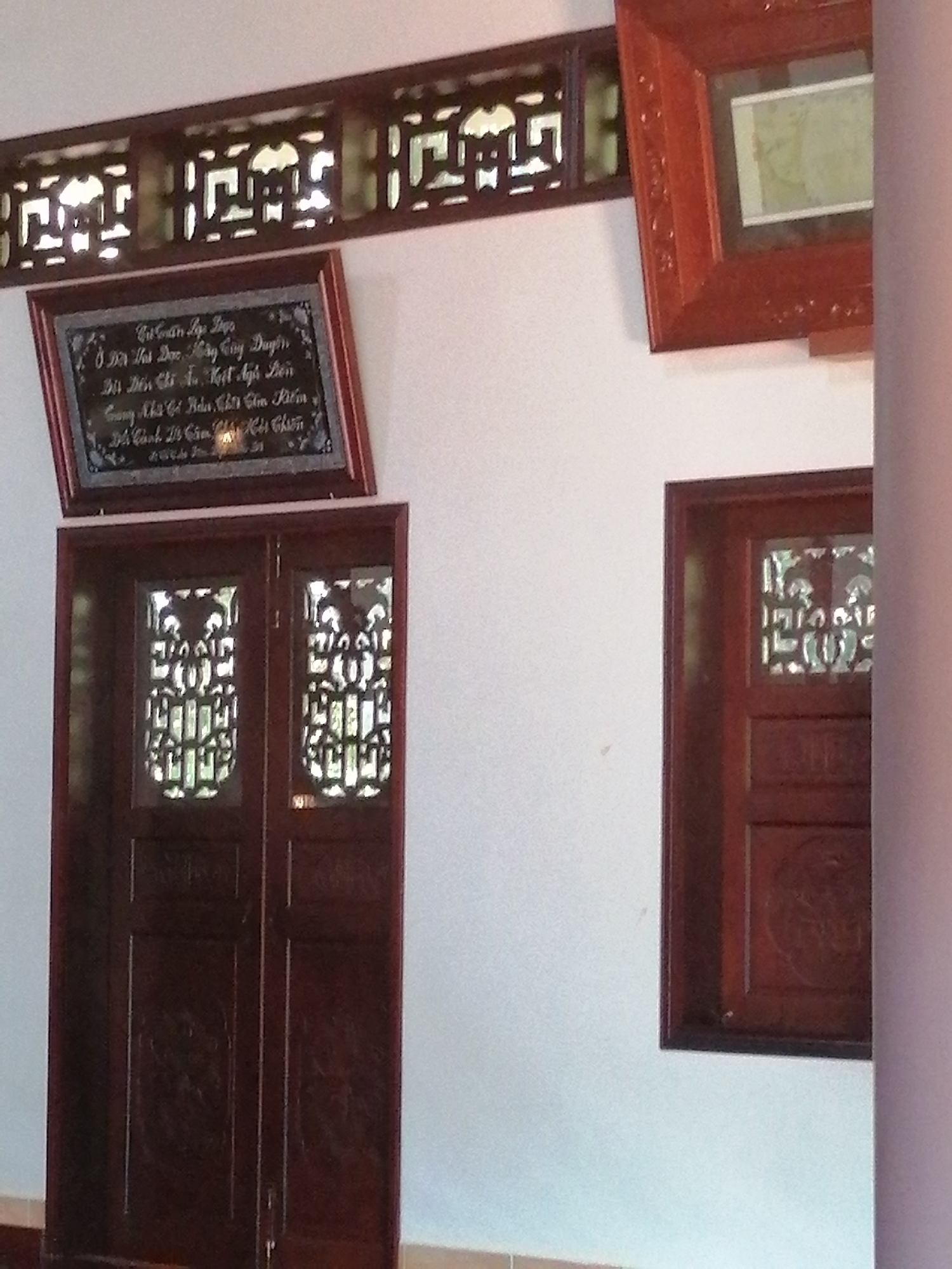 The image depicts a section of an interior wall with a large wooden door on the left and a framed window on the right. Above the door is a framed inscription, possibly a quote or a dedication. The door is dark wood with carved panels and decorative lattice work on its upper half. The window on the right is also framed in dark wood and contains a similar lattice design. Above both, a decorative wooden grate runs horizontally across the wall. The wall itself is a light, neutral color. To the far right, a portion of a pinkish-purple column is visible. The overall impression is of traditional Vietnamese architecture, likely in a historical or cultural site within Da Lat, Vietnam. No people are visible in the image, and no specific actions are being performed. The environment appears to be indoors, possibly a public building or a residence.
