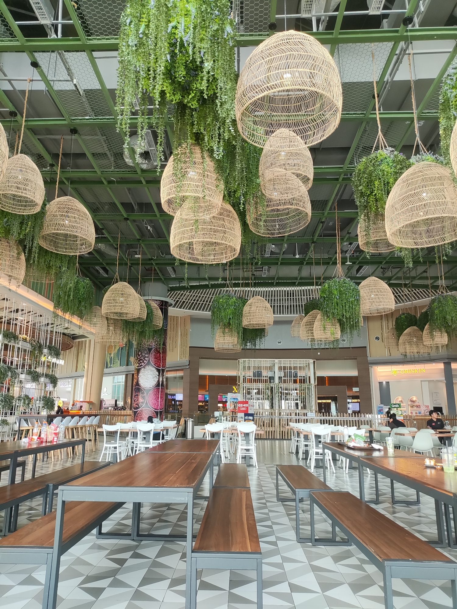 The image depicts the interior of a modern, well-lit food court or dining area with a natural, bohemian aesthetic. Large, woven pendant lights hang from the ceiling, interspersed with abundant hanging greenery, creating a canopy effect. The ceiling itself is industrial, with exposed green metal beams and ductwork, contrasting with the organic elements.
The main focus in the foreground is a row of long, wooden picnic-style tables with matching benches, all set on a patterned tiled floor. The tables and benches have dark wood tops and grey metal frames. In the mid-ground, there are more tables and chairs, including white-backed chairs and some lighter colored seating. Several tables are set with bottles of water and some food items, suggesting recent occupation.
In the background, shopfronts are visible, indicating this is likely within a larger commercial complex or mall. Signs for businesses like "OZ-KO," "GRAB," and "ORISKIN" are partially visible. A few people are seated at tables in the background, appearing to be customers or patrons of the establishment. The overall ambiance is casual and inviting, designed to create a relaxing dining experience. The location is identified as Lambangjaya, Indonesia.