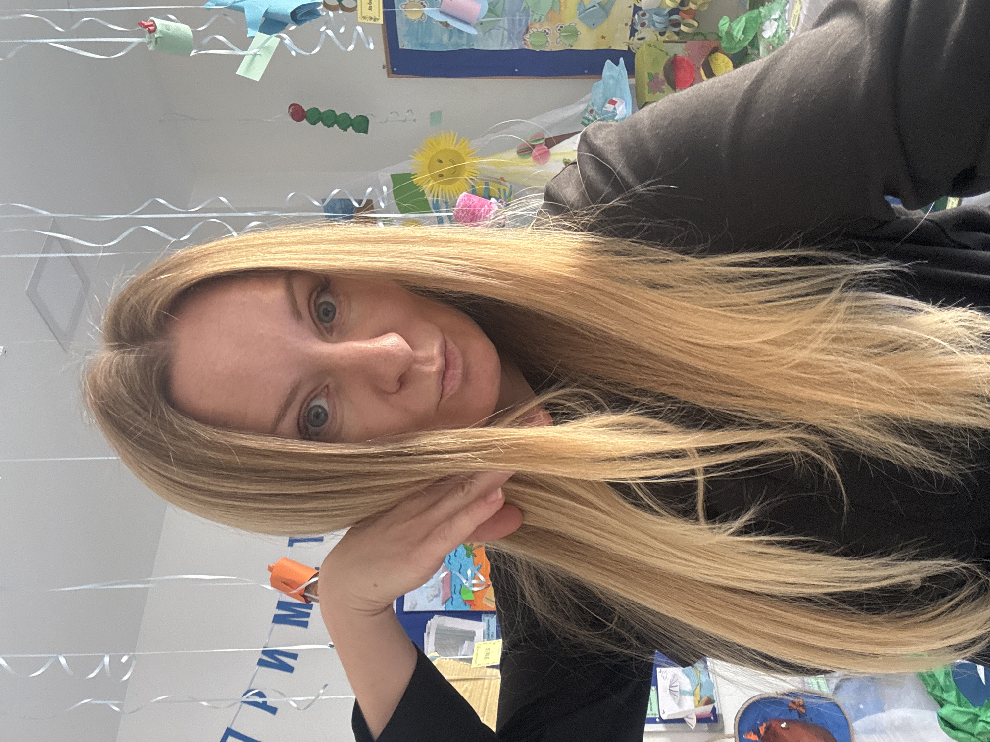A woman with long, blonde hair is in a room with decorations on the wall and hanging from the ceiling. She is wearing a black shirt and her right hand is near her face. The decorations include colorful paper crafts, artwork, and streamers. A poster with Cyrillic writing is visible. There is no indication of the city of Budva, Montenegro in the picture.