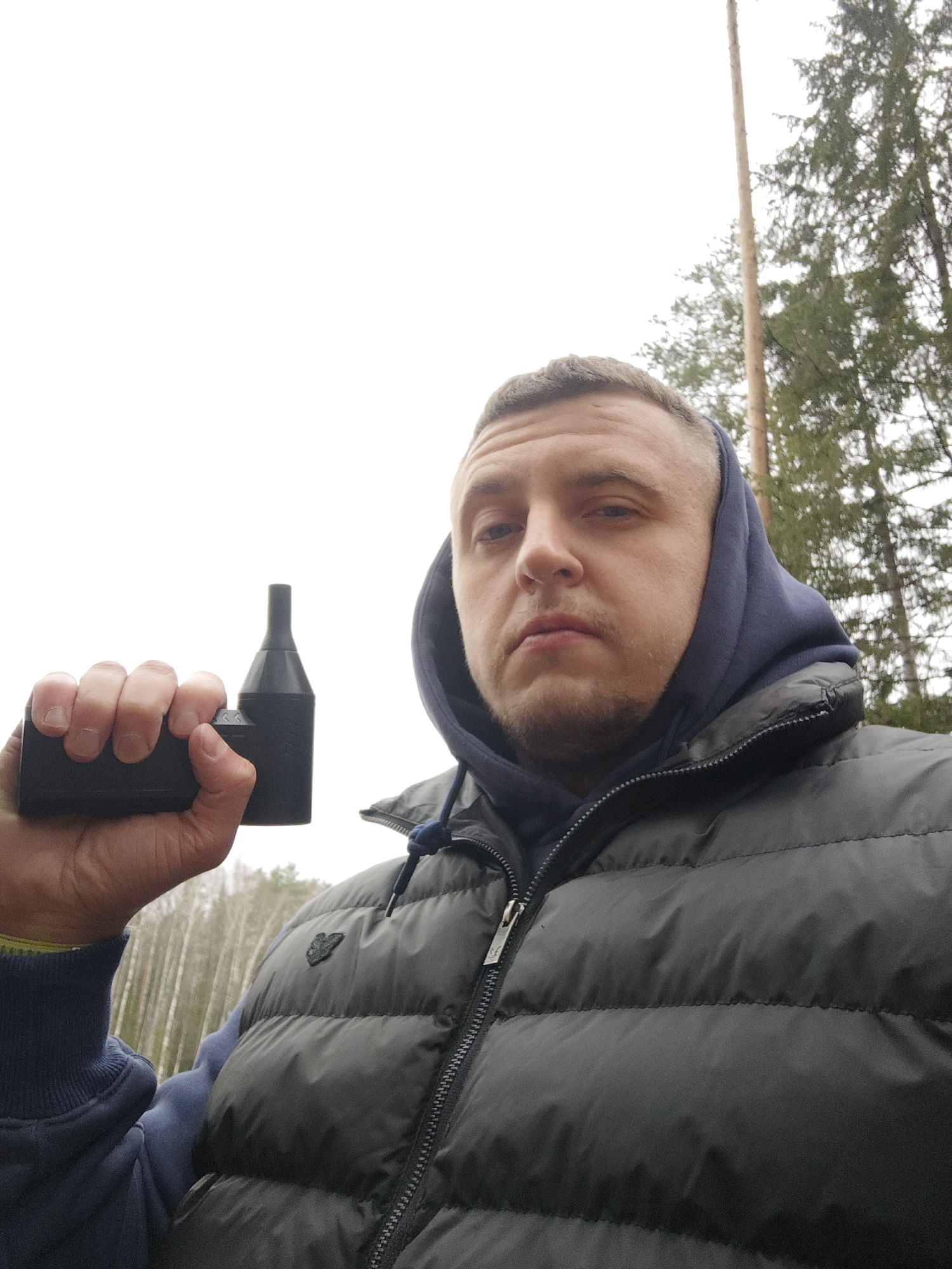 A man is featured in a close-up selfie, taken outdoors in a wooded environment. He appears to be in his 20s or 30s, with short hair and light stubble on his face, looking directly at the camera with a neutral to slightly serious expression.
He is dressed in a dark blue hooded sweatshirt, with the hood pulled up, worn under a dark grey or black quilted puffer vest. The vest has a visible front zipper and a small, dark emblem on the left chest area, the details of which are indistinct.
In his left hand, the man holds a black electronic device, which has a rectangular base and tapers upwards to a narrower, cylindrical mouthpiece. The device has some subtle texture or branding, but no specific text is legible.
The background consists of a dense arrangement of trees, including tall conifers and some bare deciduous trees, indicating a forest or woodland setting. The sky is bright and uniformly overcast, suggesting a cloudy daytime. The location is specified as Мишкино, Russia.