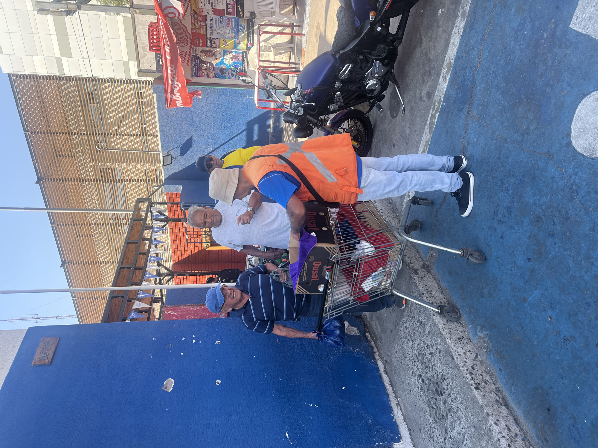 Three elderly men stand near a parked motorcycle in an urban setting, seemingly in Arica, Chile, indicated by the "Dusal" box in the shopping cart. One man in a striped shirt and blue cap stands next to a shopping cart. Another man wears a white t-shirt and a neutral-colored hat. A third man is visible in an orange vest. The environment includes a building with a blue exterior and a structure with a slat-covered awning overhead. Advertisements are visible in the background. The ground is painted blue and gray.